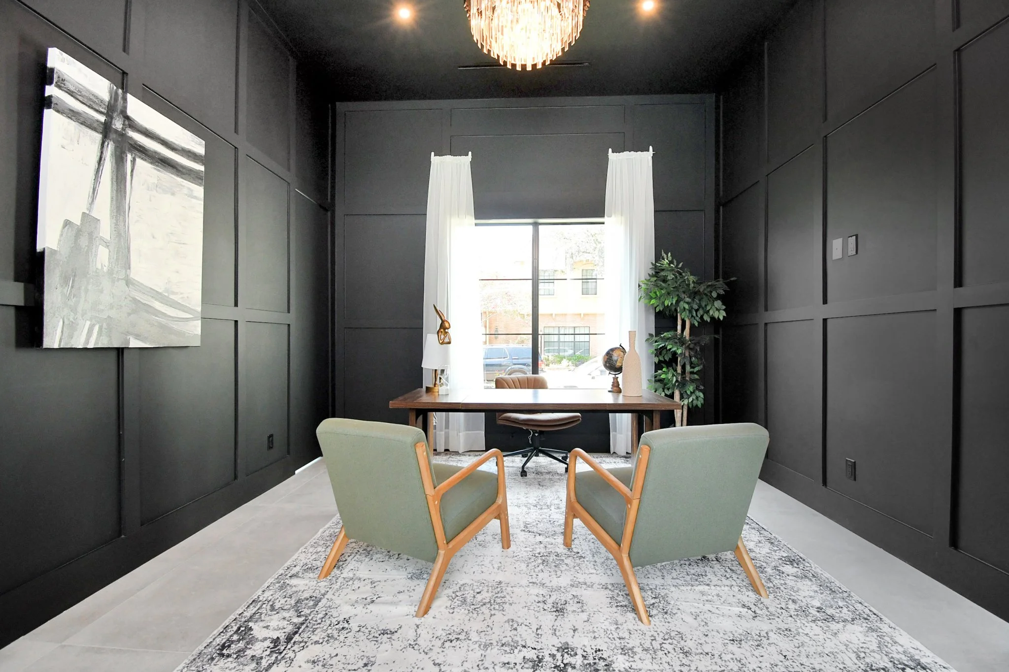 Modern black-walled office with two green armchairs facing a wooden desk, white curtains on window, indoor plant, artwork on wall, and decorative objects on desk.