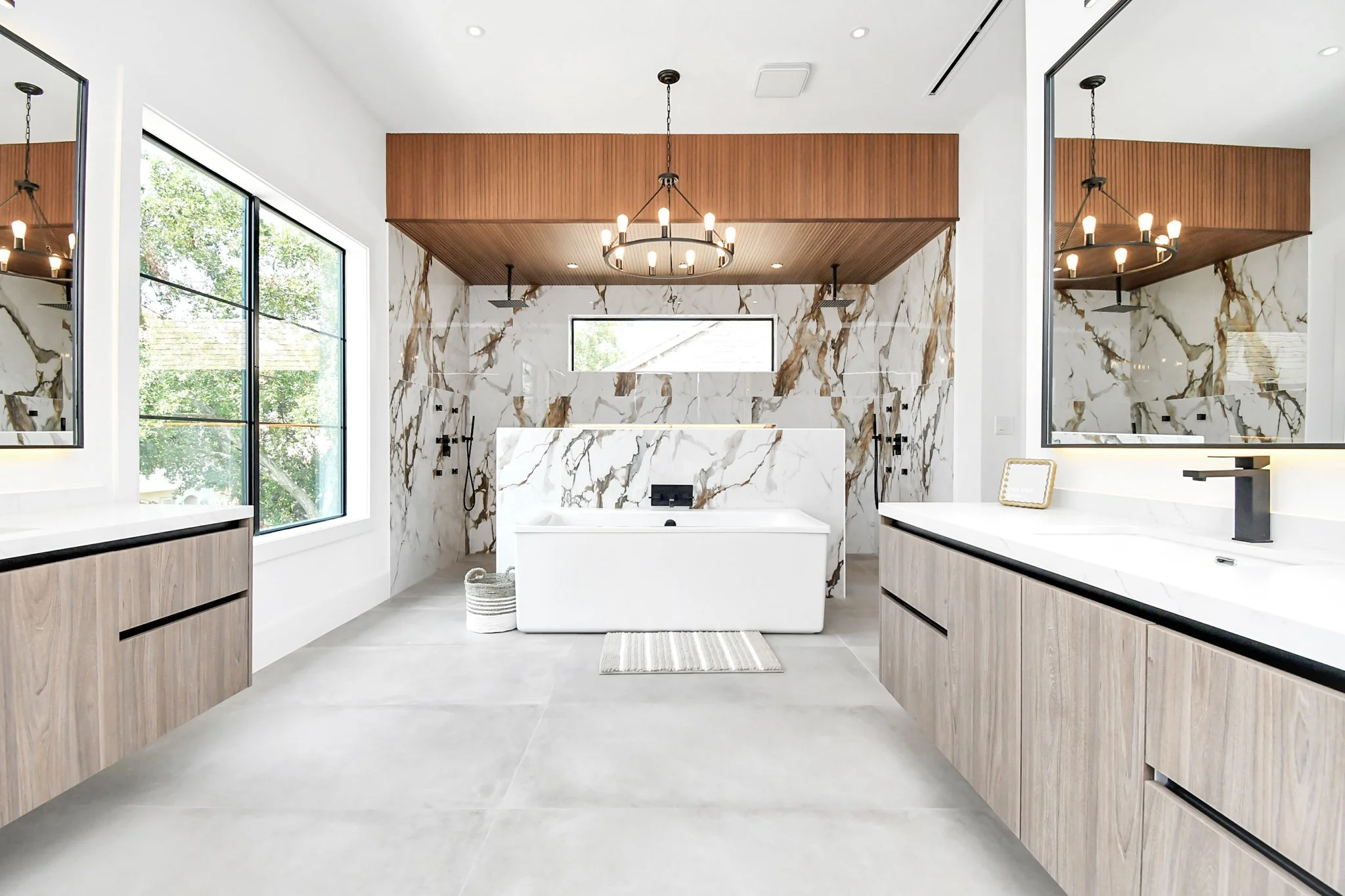 Modern bathroom with a freestanding bathtub in front of a marble accent wall, wood cabinetry, large windows, and a chandelier.