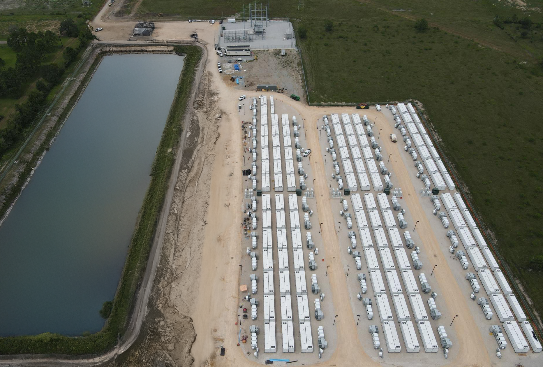 Aerial view of a large outdoor location with rows of white energy storage laid out on a wide dirt area, adjacent to a body of water on the left and green fields on the right.