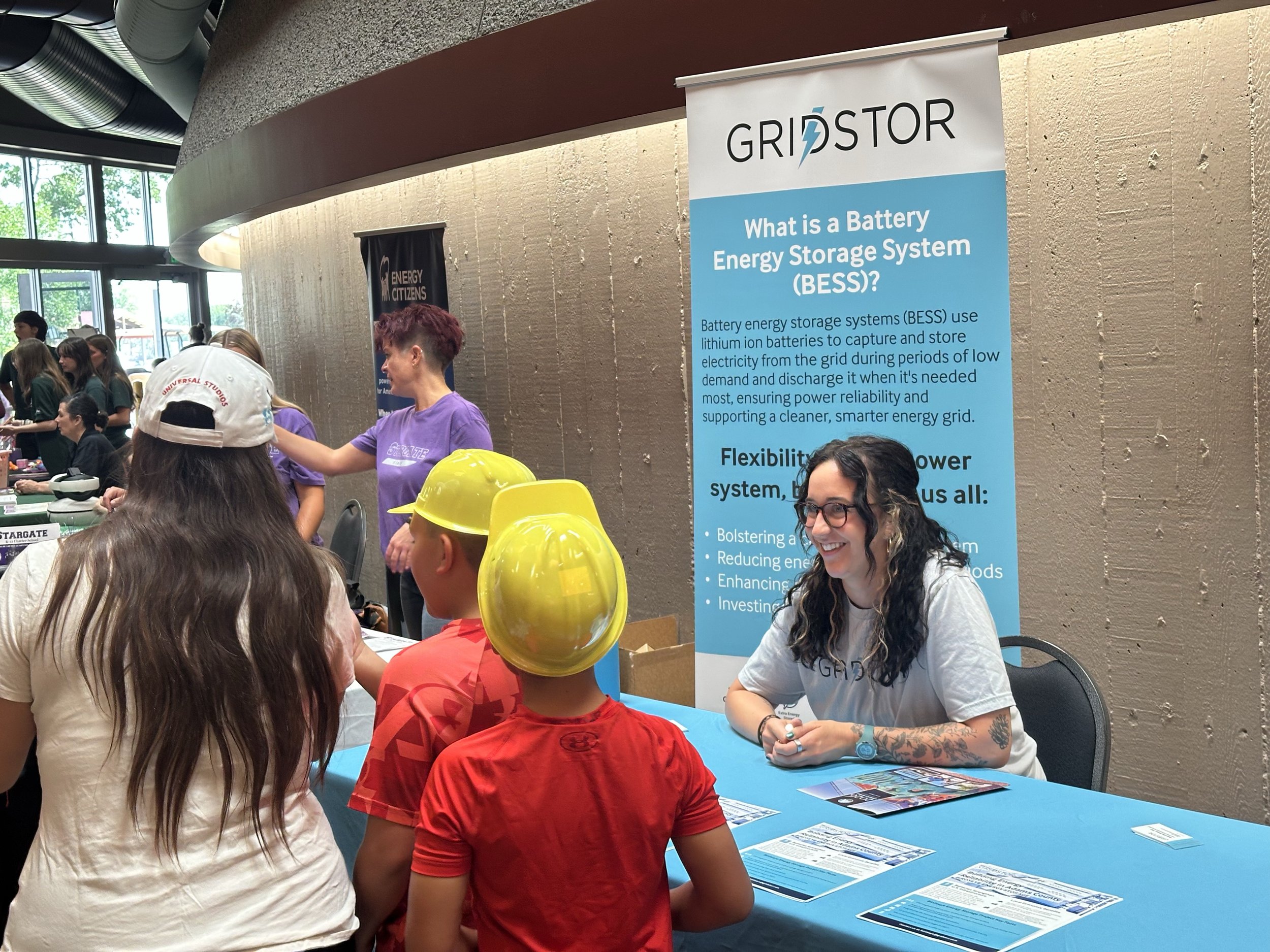 People gathered at an indoor event around a booth with a blue banner titled 'GRIDSTOR' explaining battery energy storage systems, with tables and pamphlets, and some individuals wearing yellow hard hats.