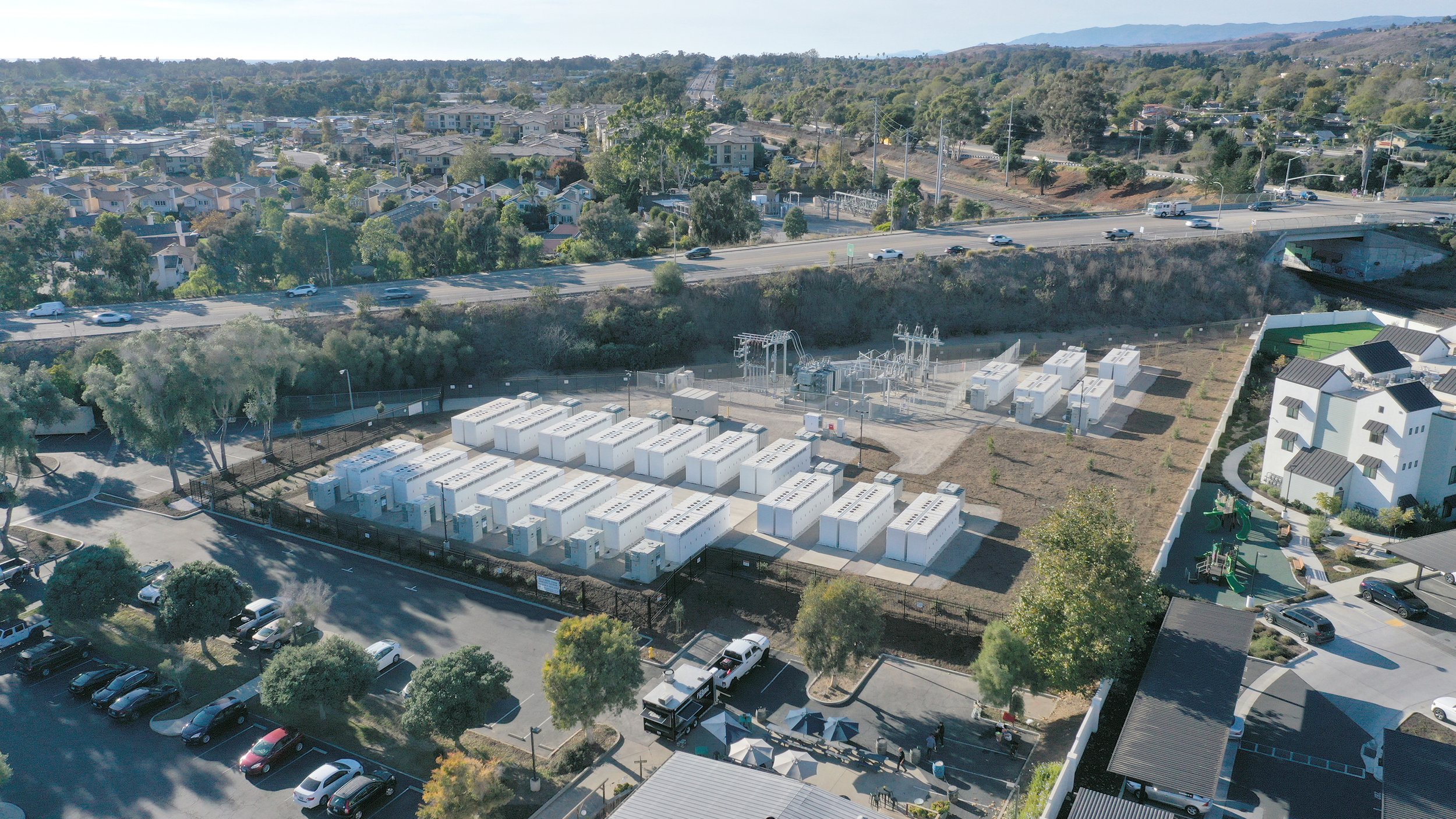 Aerial view of a neighborhood with residential houses, a highway with moving cars, and a large utility substation with multiple white transformers and electrical equipment.