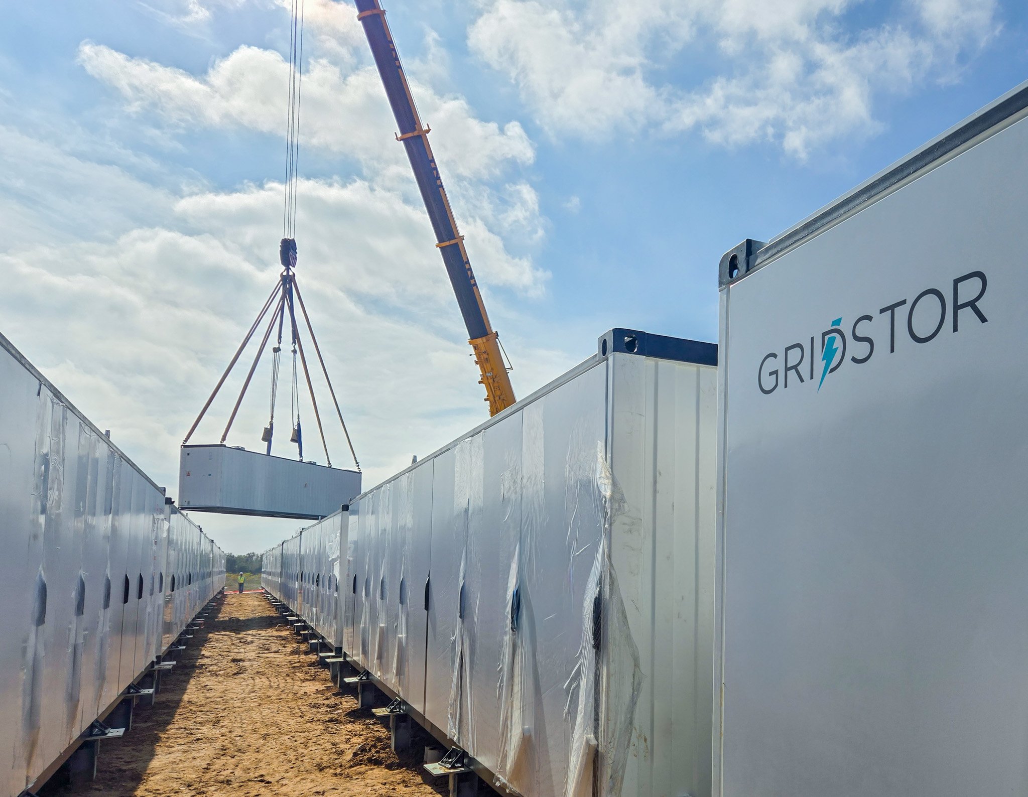 View of Large white metal energy storage containers in rows, with a crane lifting one in the background, under a partly cloudy sky.