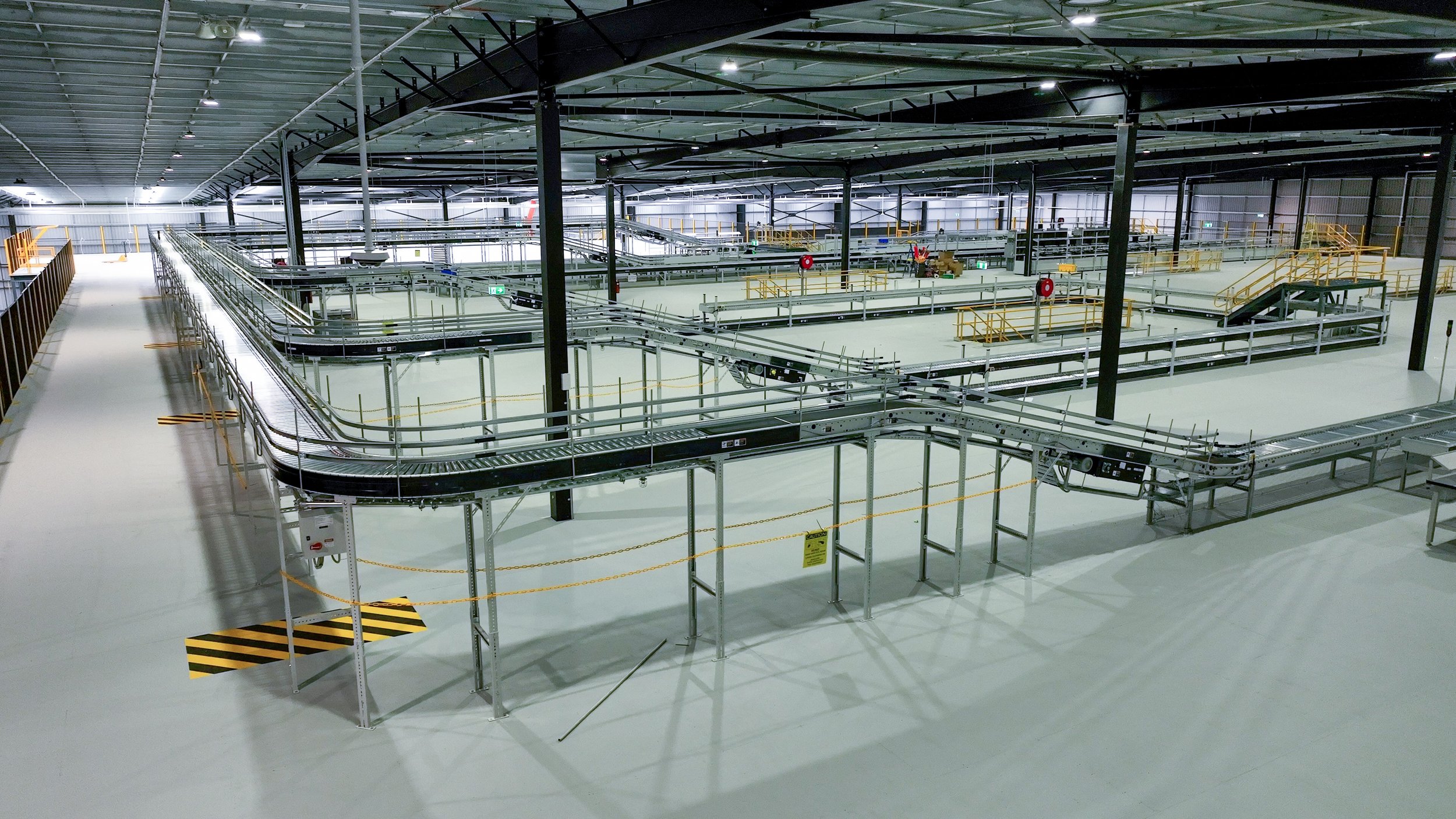 Interior view of an empty warehouse or manufacturing facility with metal conveyor belts, stairs, and safety barriers.
