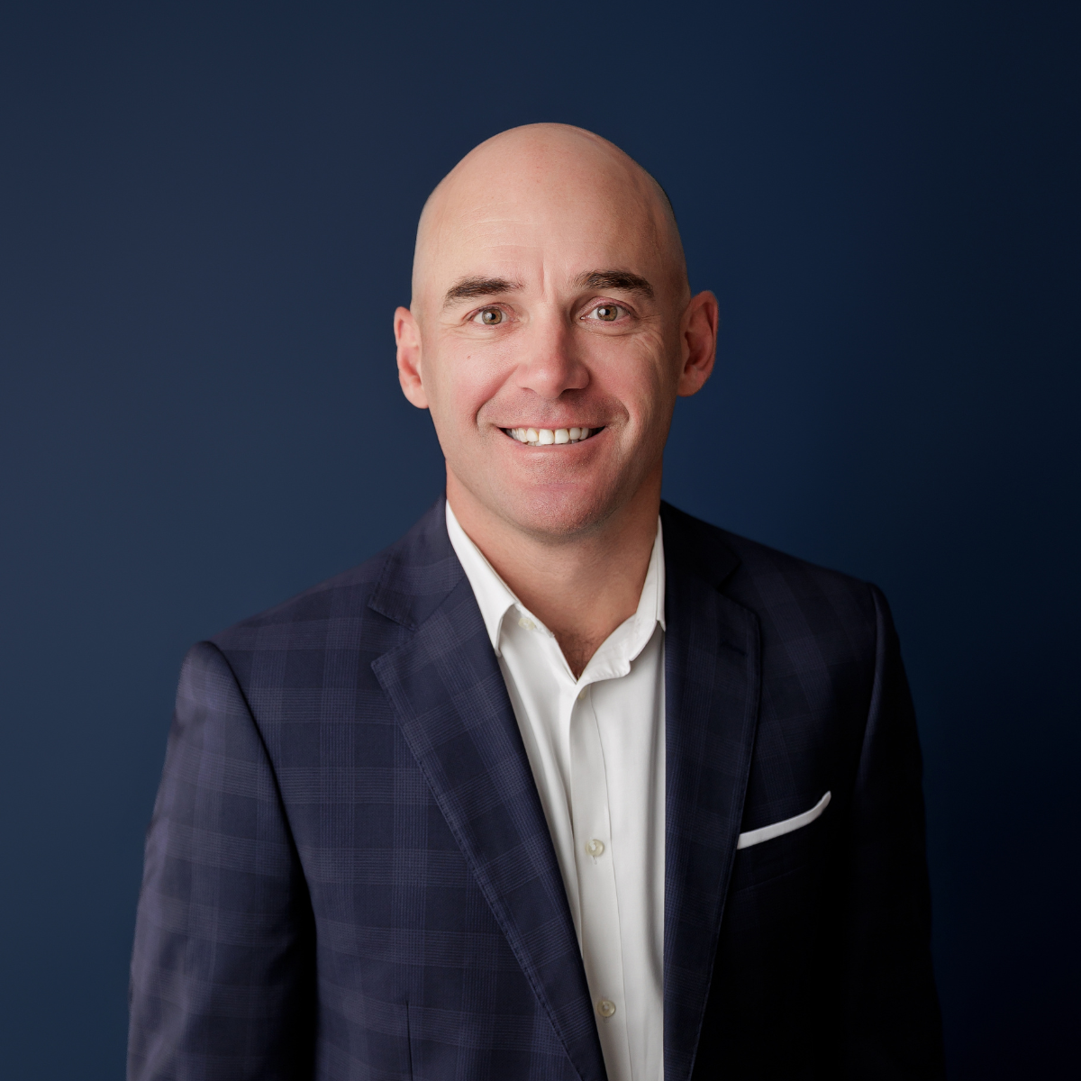 Headshot of a smiling bald man wearing a navy checkered suit and white dress shirt against a dark blue background.