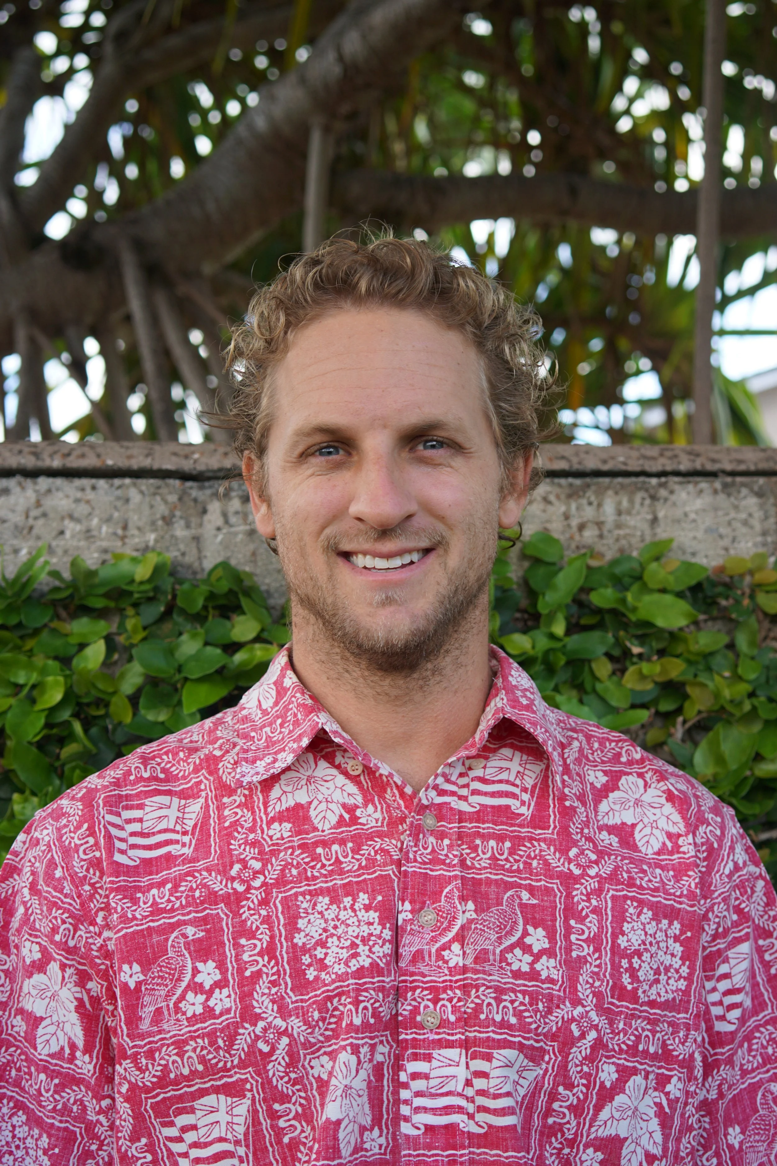 Derek Risch standing in front of a lush green leafy background with a tree above.