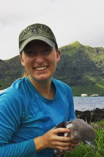Jessica Idle holding a small bird near water with mountains and green countryside in the background.