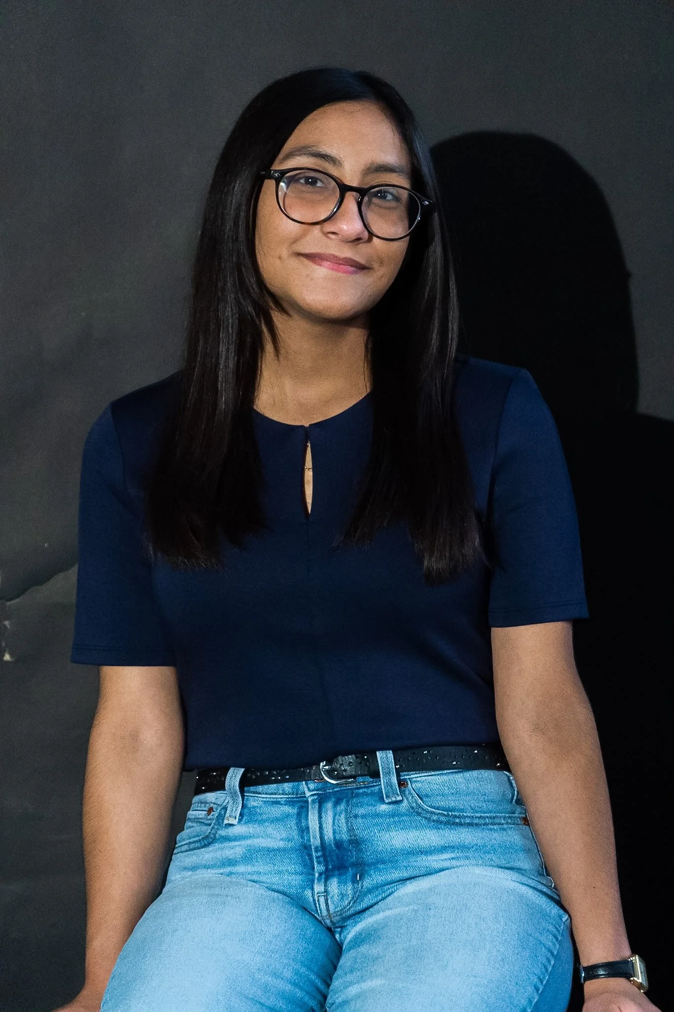 A young woman with long black hair, glasses, and a smile, wearing a navy blue top and light blue jeans, sitting against a dark background.