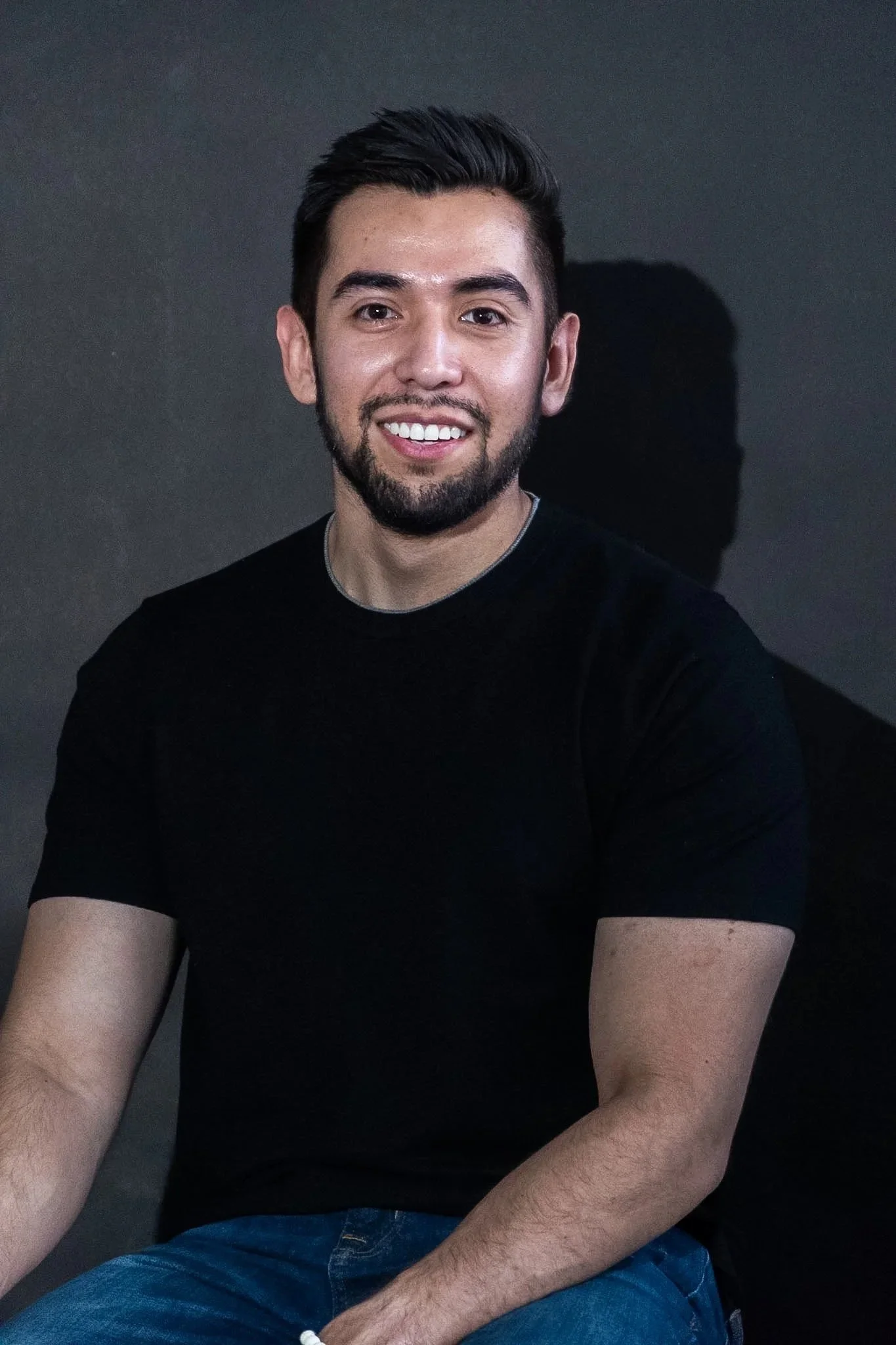 A young man with dark hair and facial hair smiling, wearing a black t-shirt, sitting in front of a dark background.
