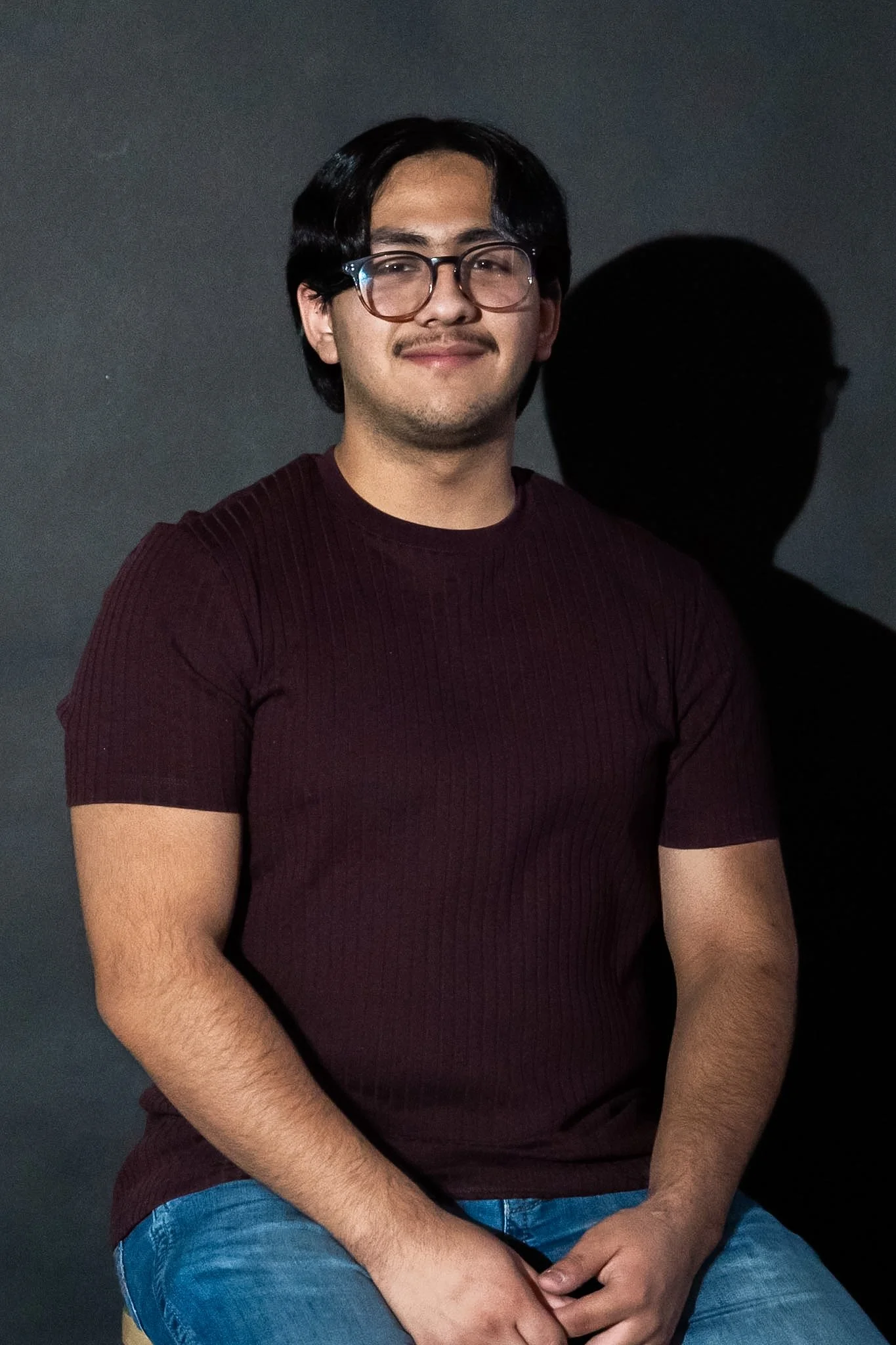 A young man with black hair, glasses, and a light mustache and beard, wearing a maroon ribbed shirt and blue jeans, sitting against a dark background with a shadow cast to his right.