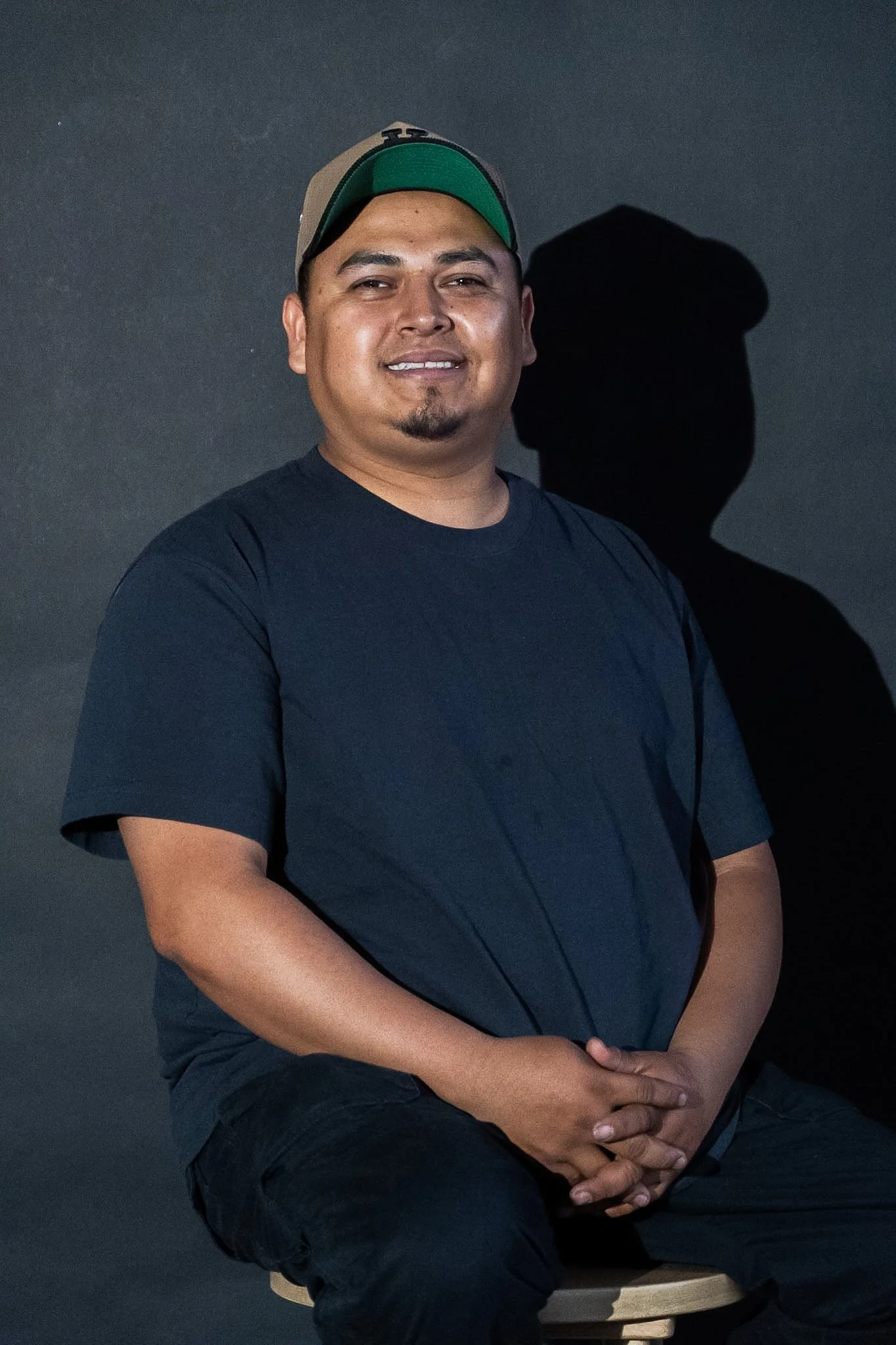 A man wearing a beige and green cap, a black t-shirt, and black pants, sitting on a stool against a dark background, smiling. His shadow cast on the wall behind him.