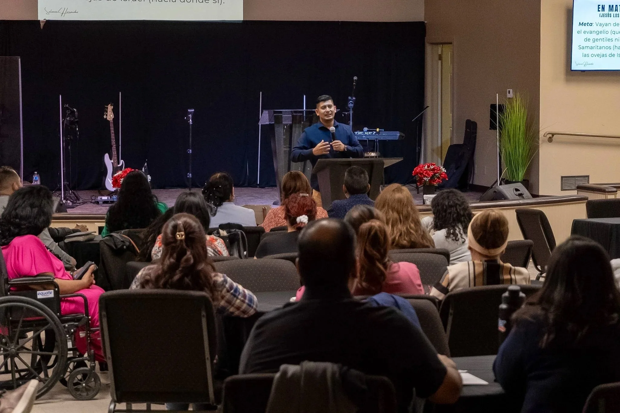 A man speaking at a podium during a presentation or church service, with a seated audience in front of him, some taking notes or listening attentively, and a stage with musical instruments in the background.