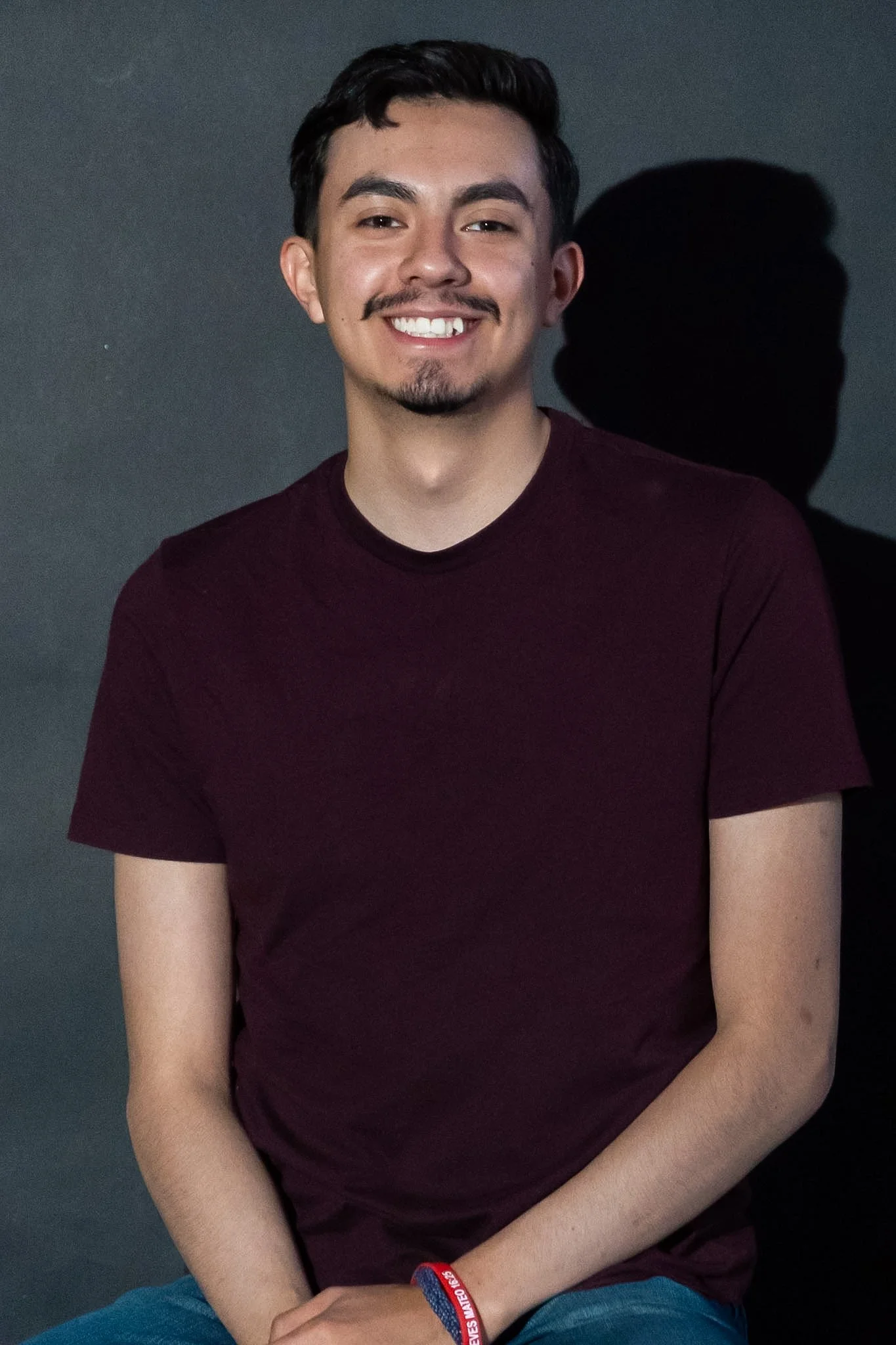 Young man with dark hair, mustache, and goatee, smiling, wearing a maroon t-shirt and a red wristband, sitting against a dark background with a shadow behind him.