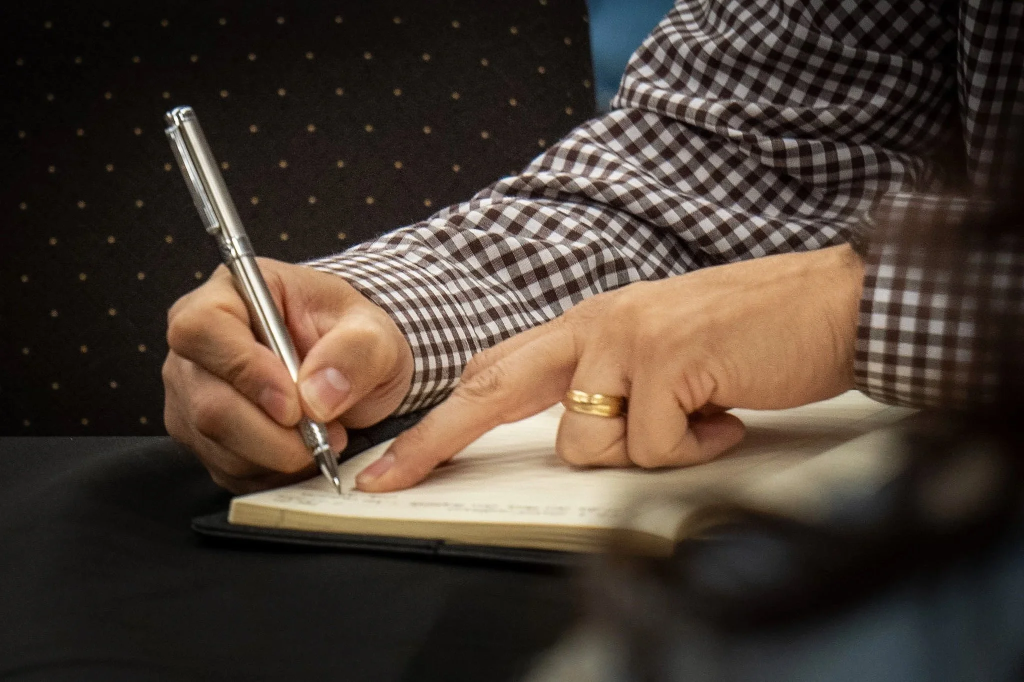 A person in a checkered shirt writing in a notebook with a silver pen.