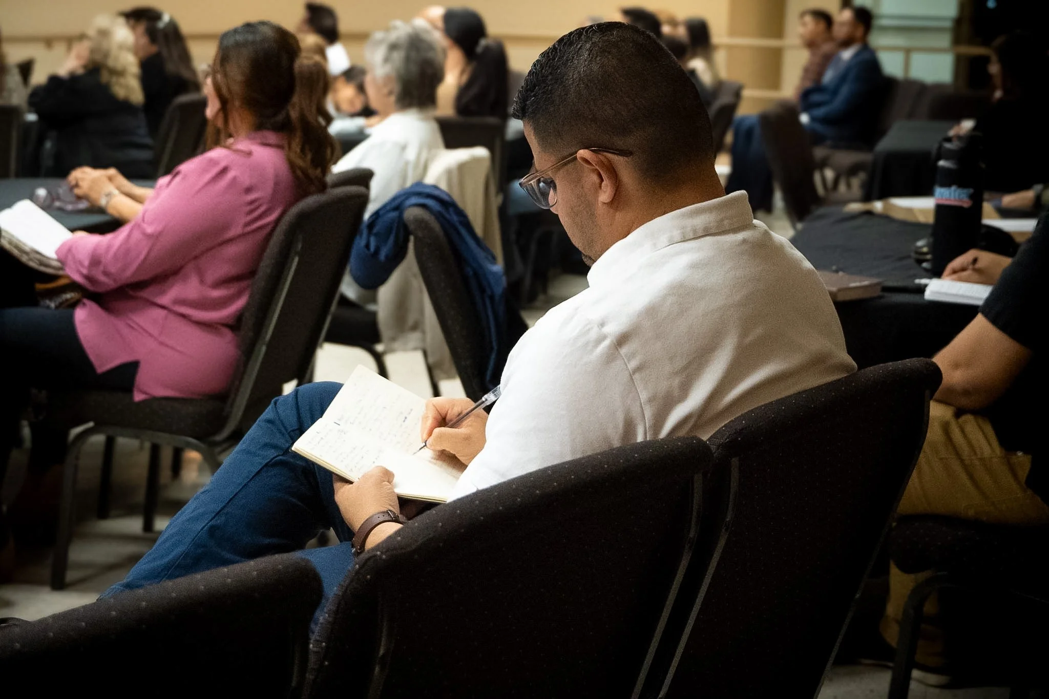 A man with glasses and a white shirt sitting in a conference room, taking notes in a notebook during a meeting or seminar, with other attendees seated around him.