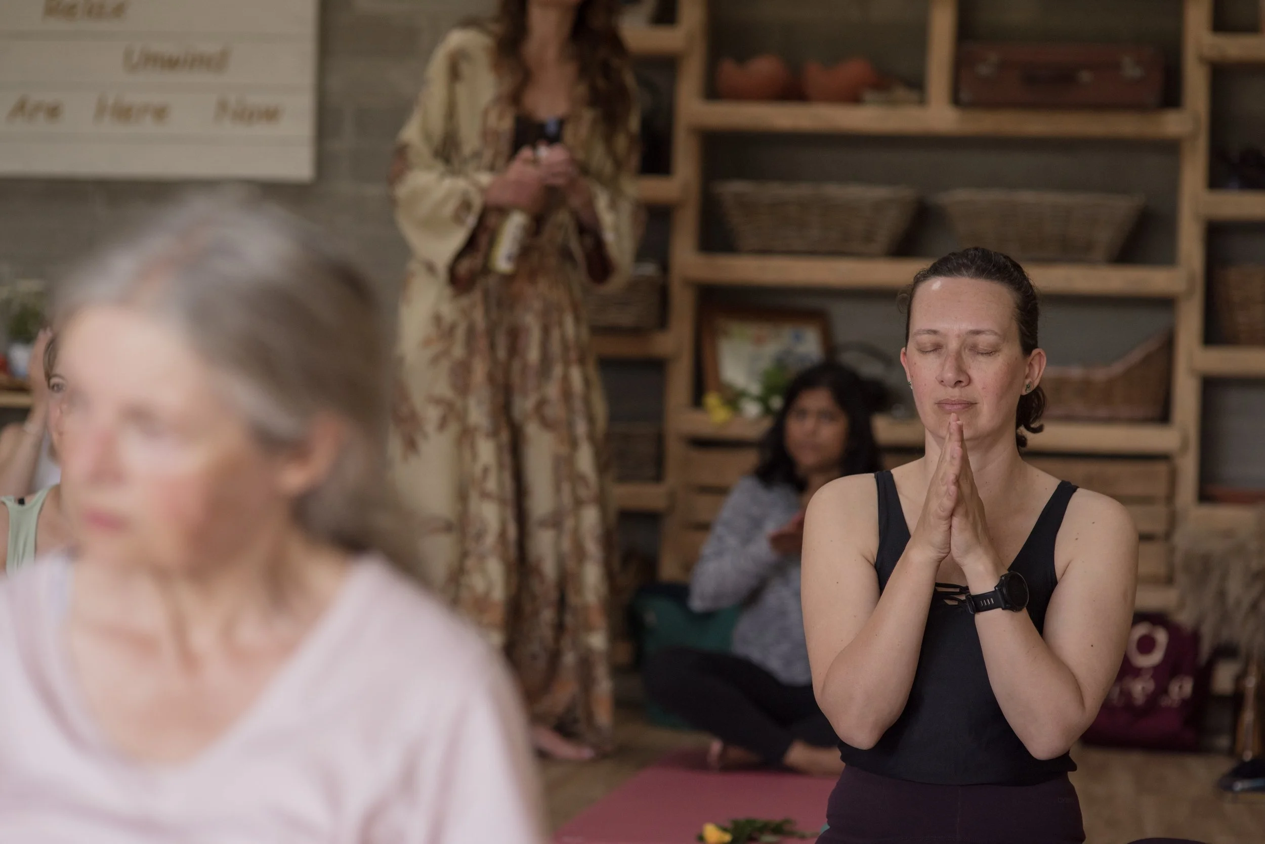 Woman with hands bin prayer, preparing for meditation and two women in the background faded seated on yoga mat