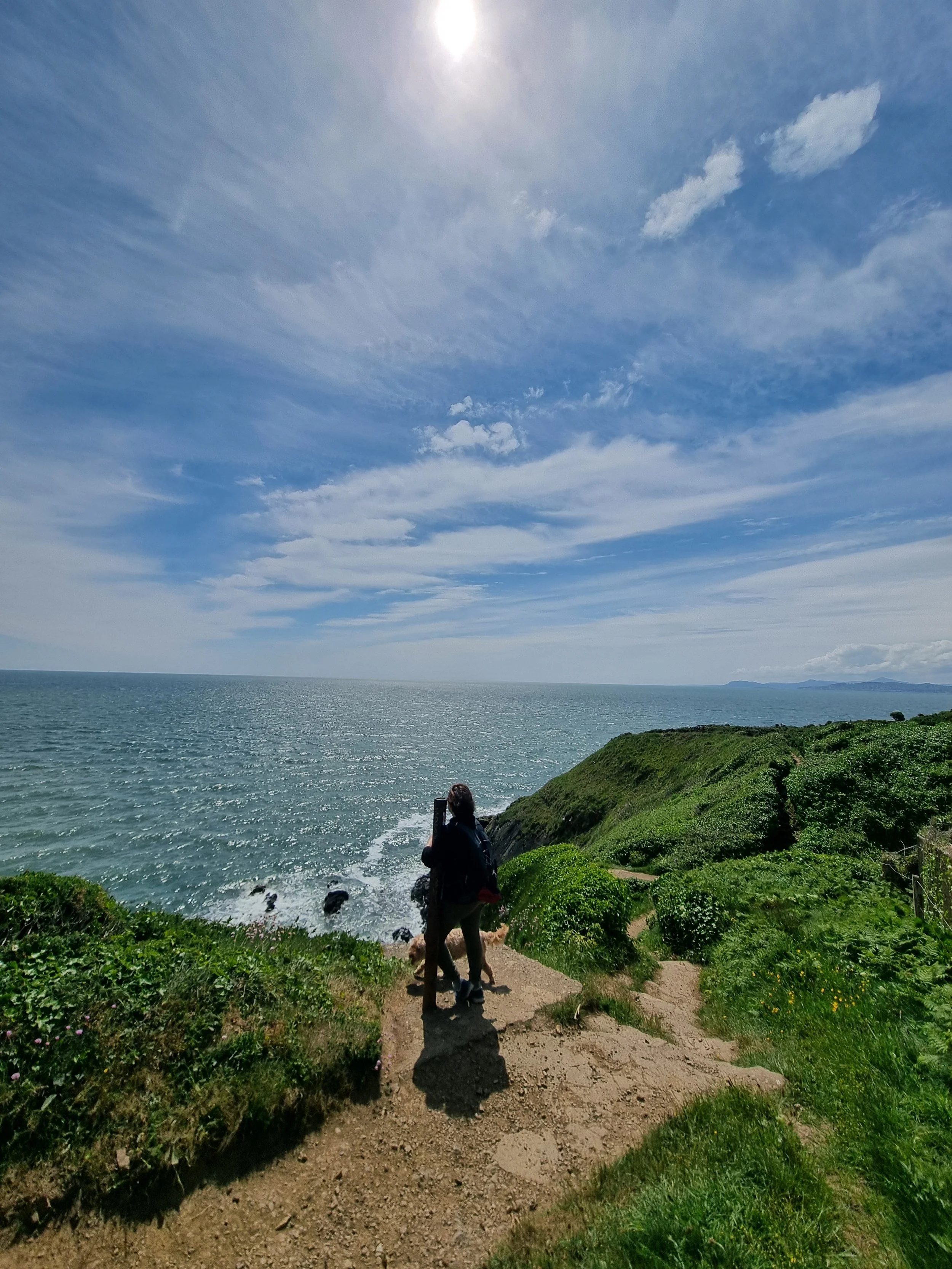 Person standing on a coastal cliff path overlooking the sea under a bright sun and cloudy sky.