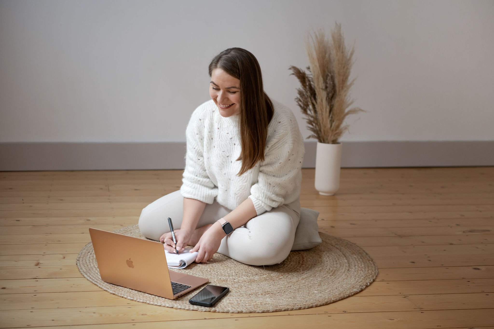 Ana Stefan sitting on rug with laptop and notepad, writing, vase with pampas grass in background.