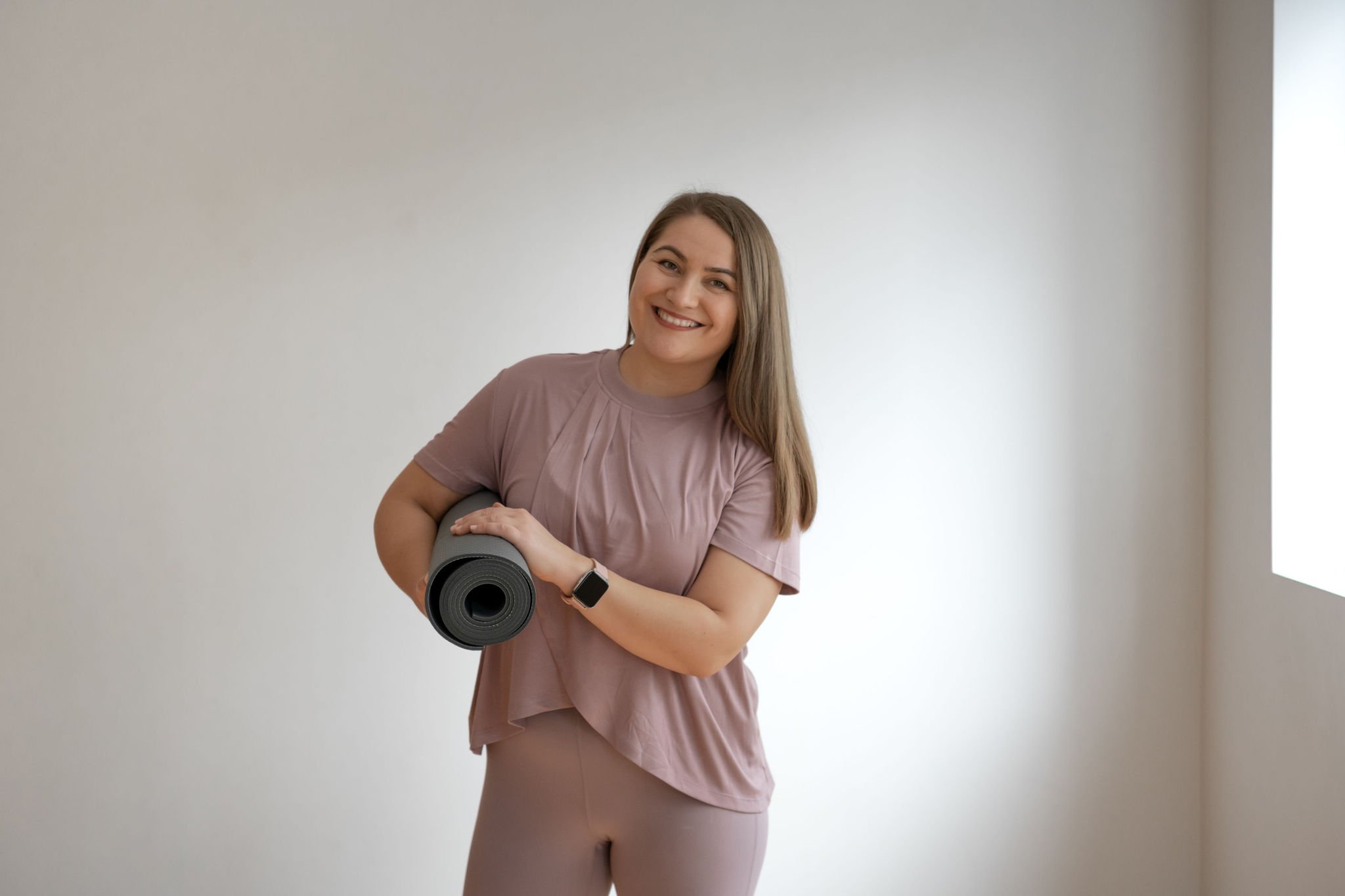 Ana holding yoga mat, wearing pink workout clothes, smiling indoors.