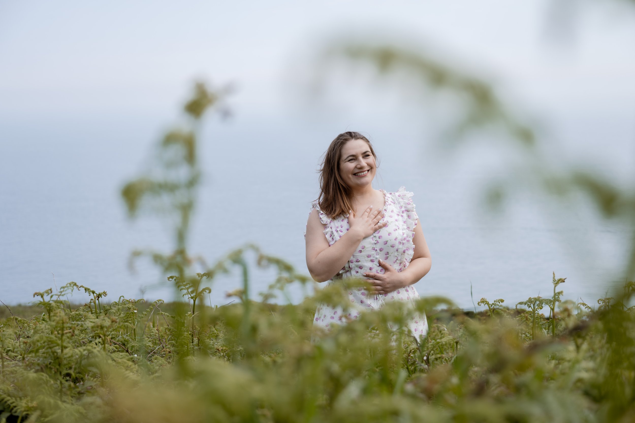 Smiling Ana in floral dress standing in field with ocean in background.