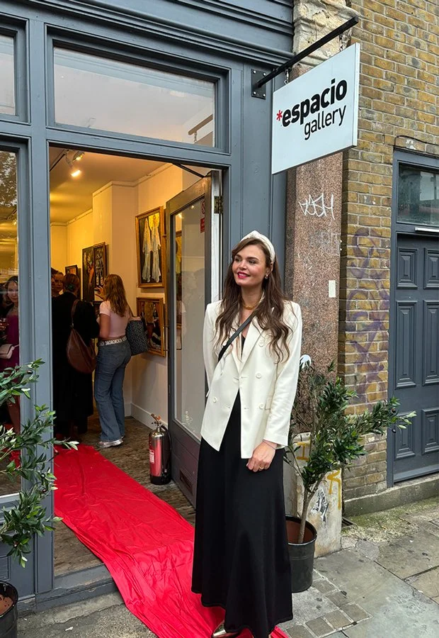 A woman in elegant attire standing outside an art gallery with a red carpet leading inside, with other visitors and artwork visible inside.