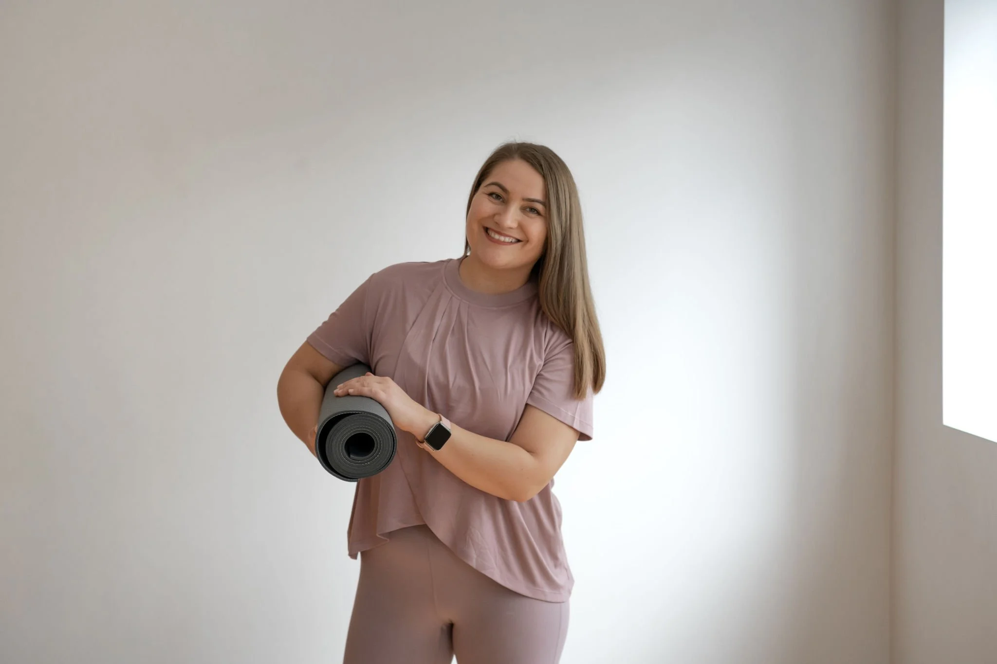 A smiling woman holding a rolled-up yoga mat in a bright room with plain white walls.