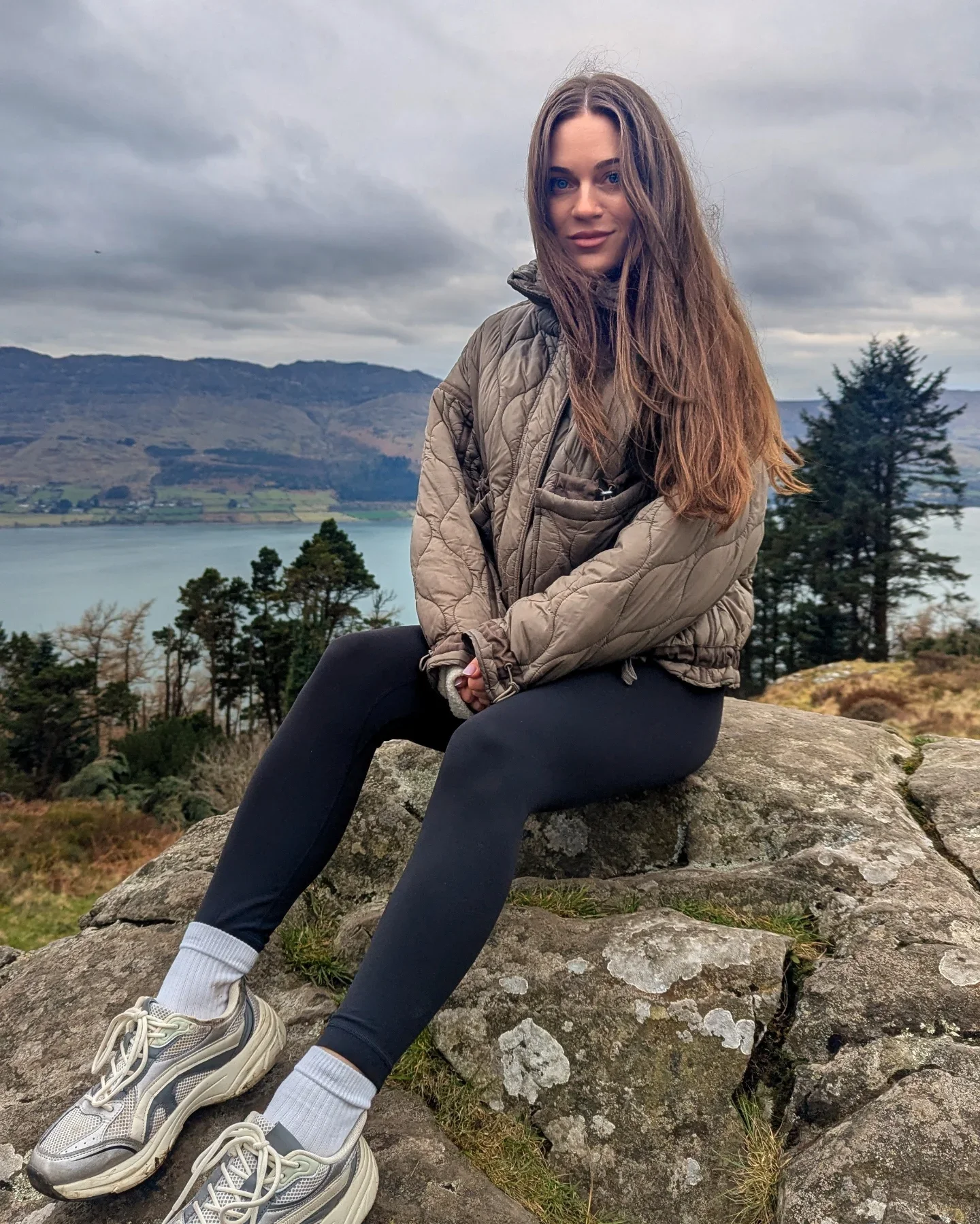 A young woman sitting on a large rock outdoors with a scenic lake, trees, and mountains in the background. She is wearing a tan quilted jacket, black leggings, white socks, and athletic shoes. Overcast sky.