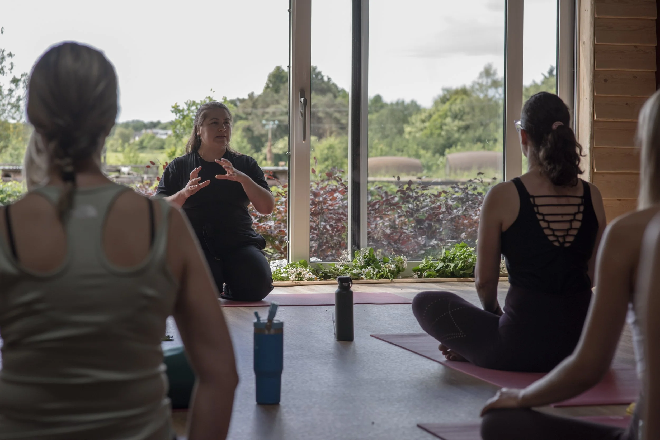 A woman leading a yoga or meditation class in a room with large windows overlooking trees. Participants are sitting on yoga mats, facing the instructor.