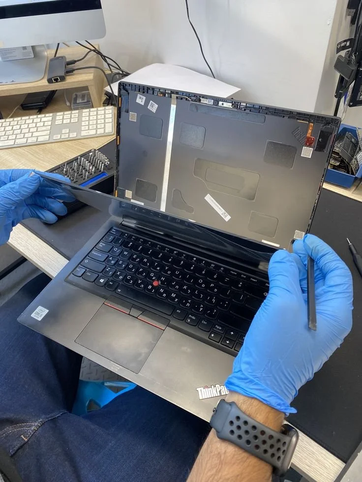 Persona reparando una laptop con la pantalla levantada, usando guantes de procedimiento, en un espacio de trabajo con herramientas y accesorios tecnológicos.