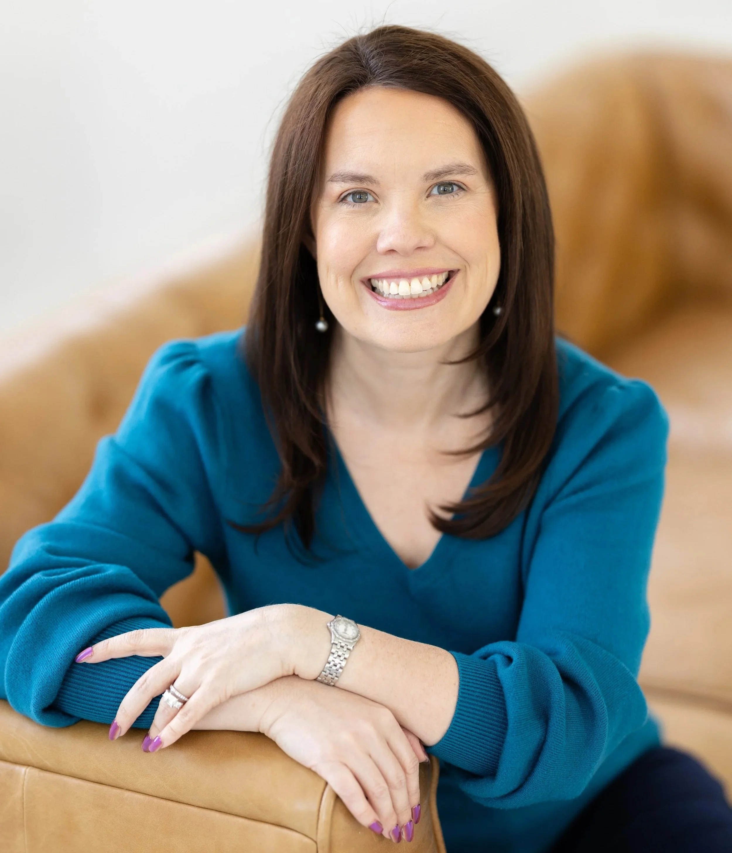 Close-up of a smiling woman with blue eyes and brown hair, wearing a blue top and jewelry, resting her arms on a tan leather surface.