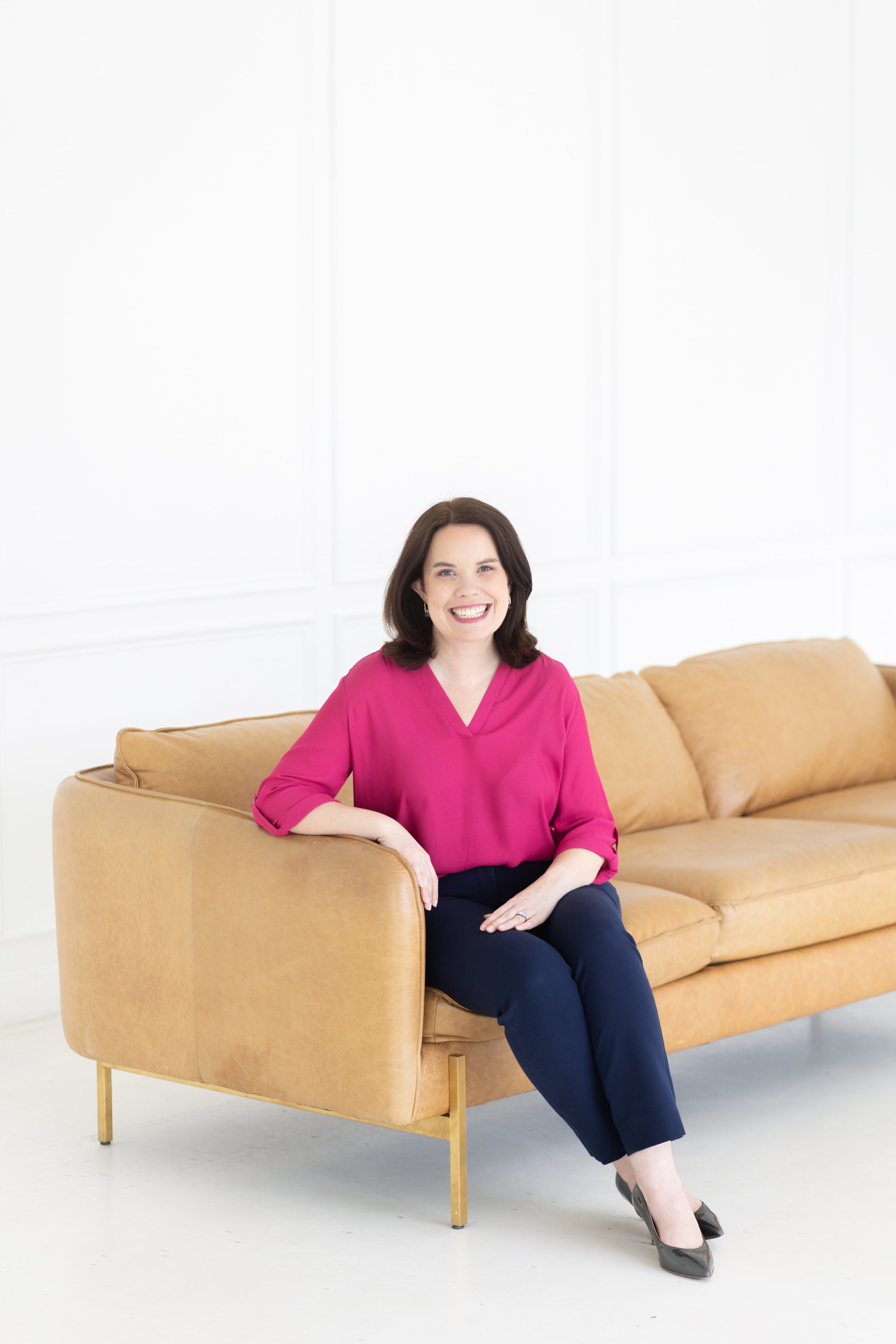 A woman with shoulder-length brown hair, wearing a bright pink blouse and dark pants, is sitting on a tan-colored sofa in a bright room with white walls, smiling at the camera.