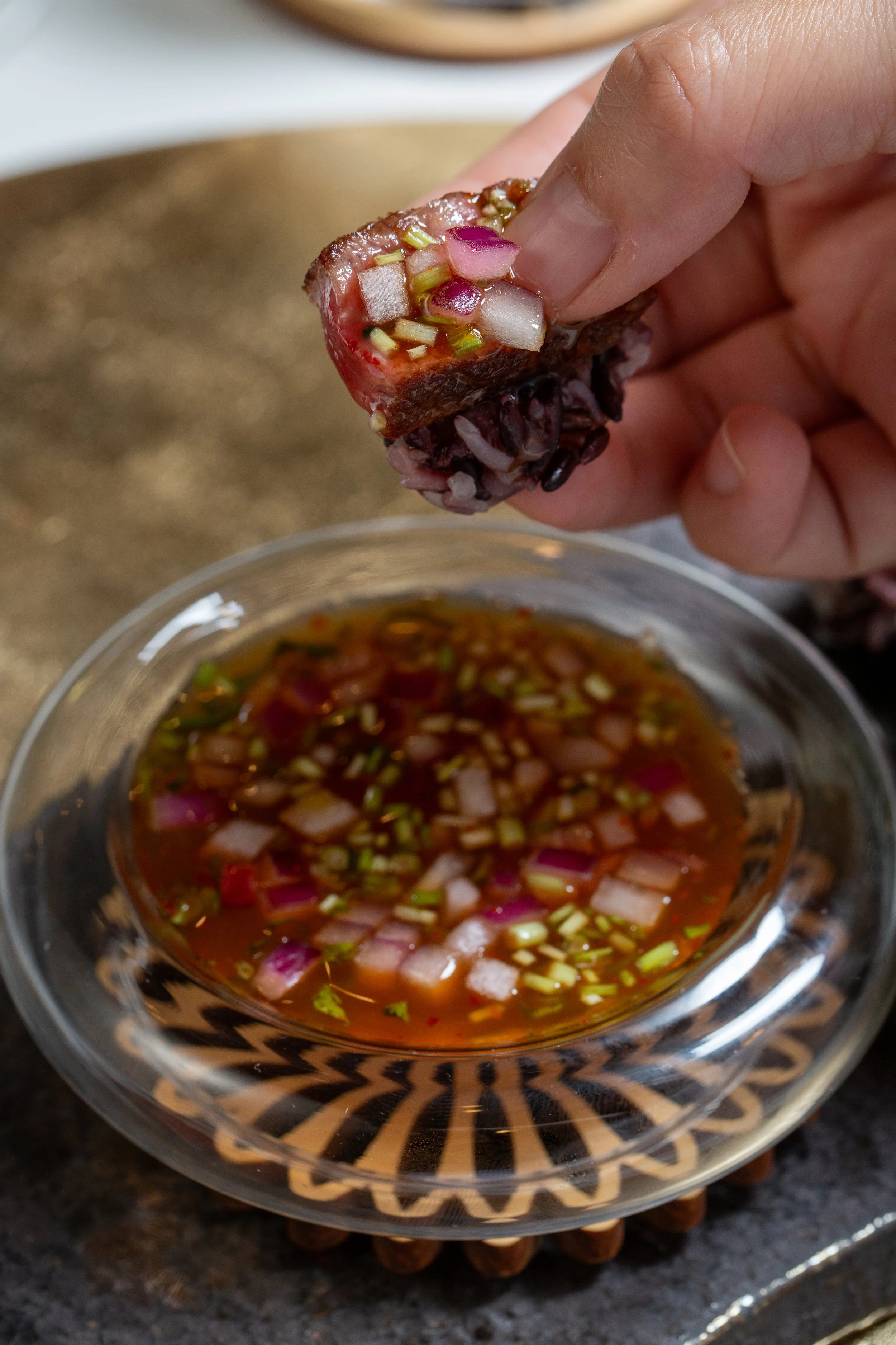 A hand holding a piece of meat topped with chopped onions over a bowl of chopped onions and sauce.