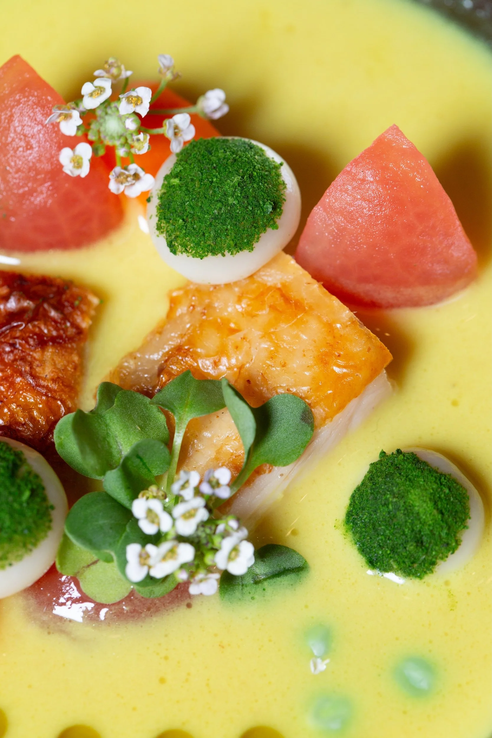 Close-up of a gourmet dish featuring sliced tomatoes, small white flowers, green herbs, and cilantro oil.