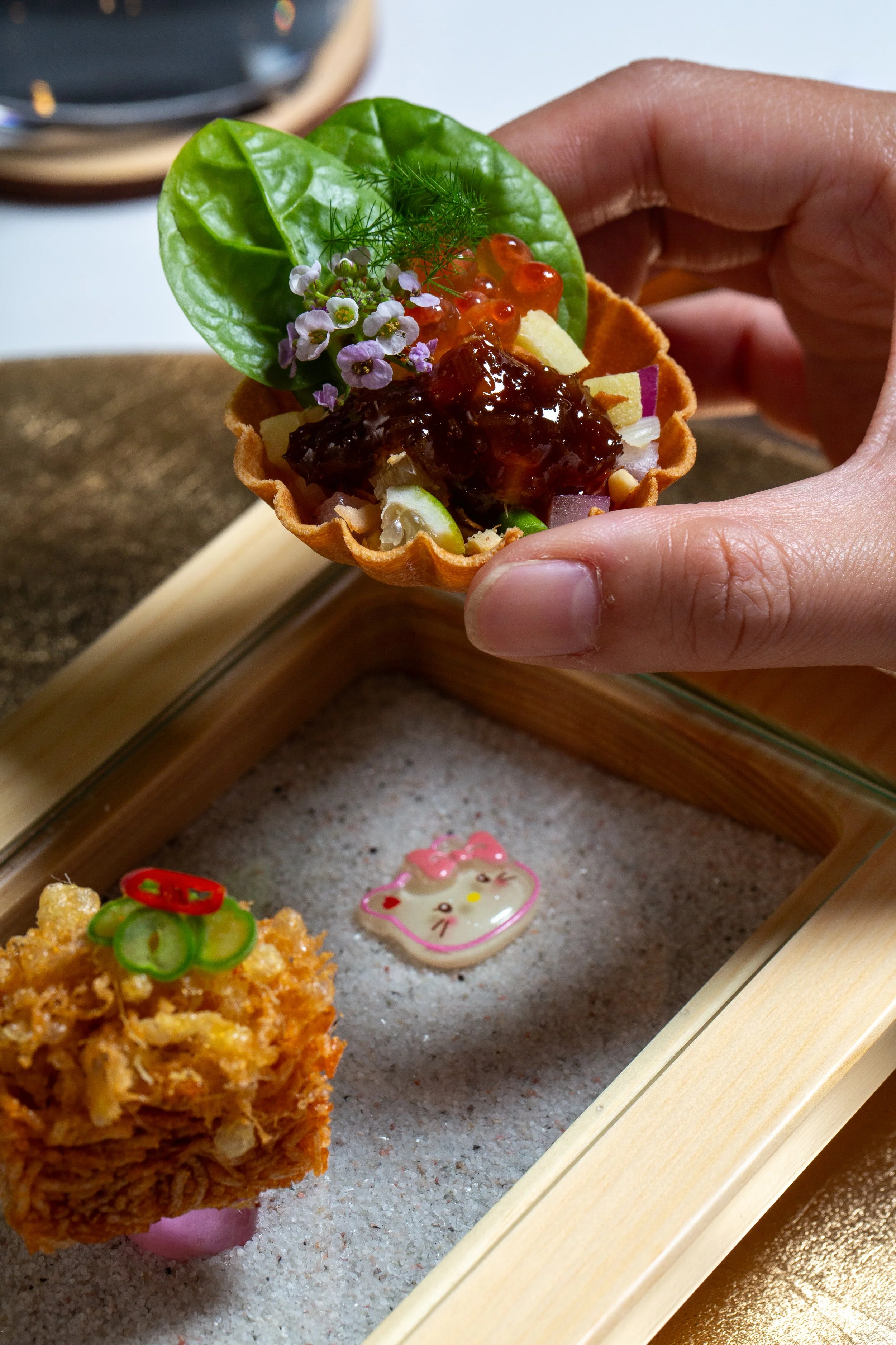 Close-up of a hand holding a small edible tart with a green leaf, purple flowers, and orange jelly beans on top, placed inside a waffle crust. Other fried food items and a cute drink umbrella decoration are visible on a tray in the background.