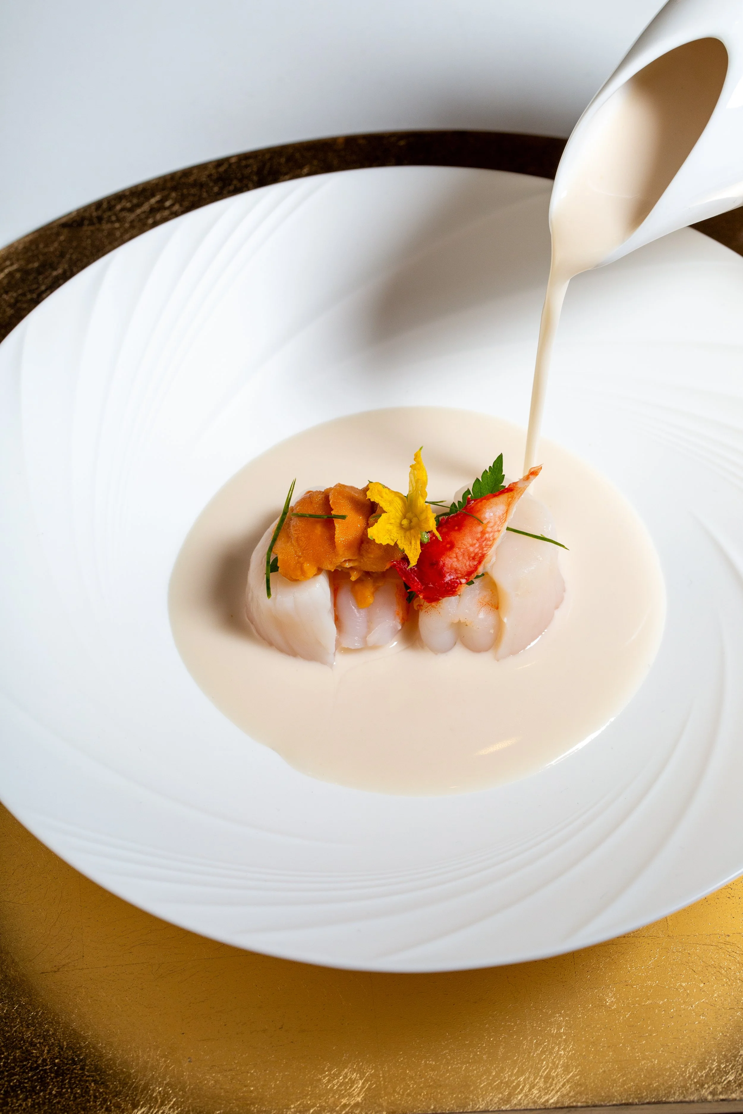 A white bowl with a dark rim contains a creamy sauce or soup, with a seafood dish featuring two large shrimp, garnished with a yellow edible flower, green herbs, and an orange relish, with white sauce being poured over it from a white spoon.
