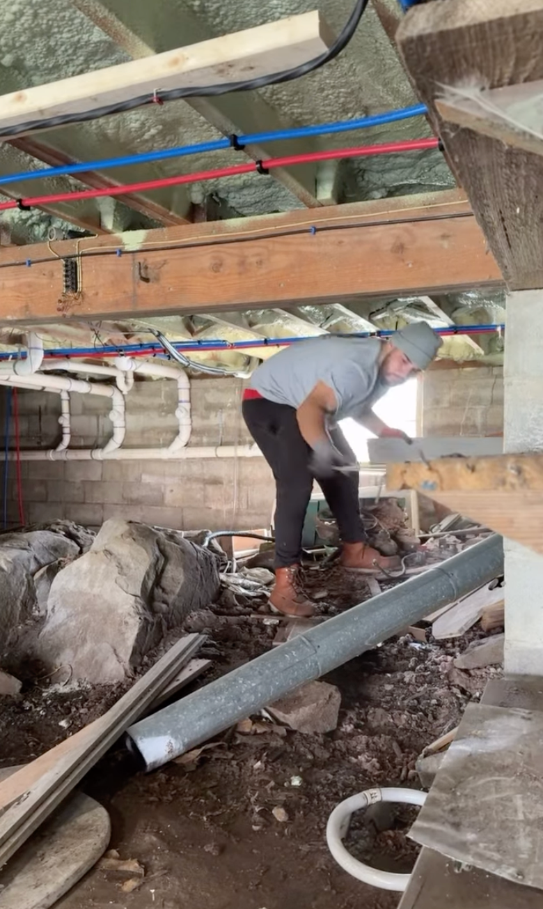 A construction worker wearing a gray shirt, black pants, brown boots, and a gray beanie is working on a construction site, using a tool on the floor surrounded by rocks, pipes, and construction debris.