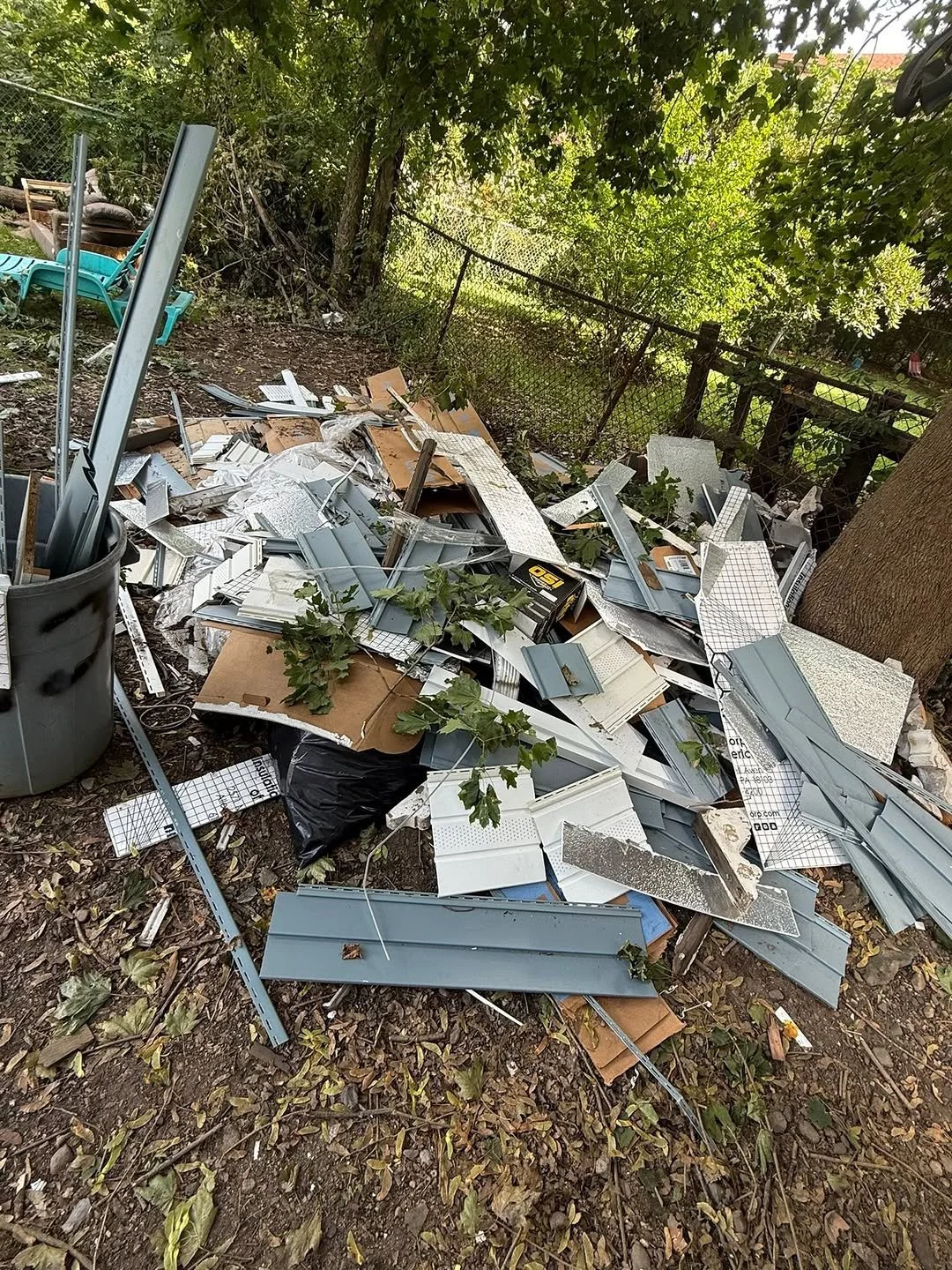 Pile of torn and discarded construction or electrical materials, including metal and plastic components, on the ground in a backyard with trees and a fence.