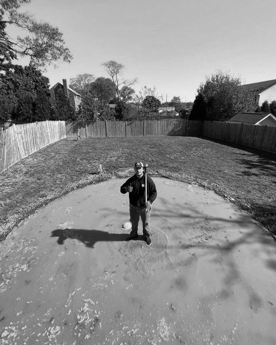 A man standing on a construction site holding a tool, with a partially completed concrete surface and a wooden fence surrounding the area in a backyard.