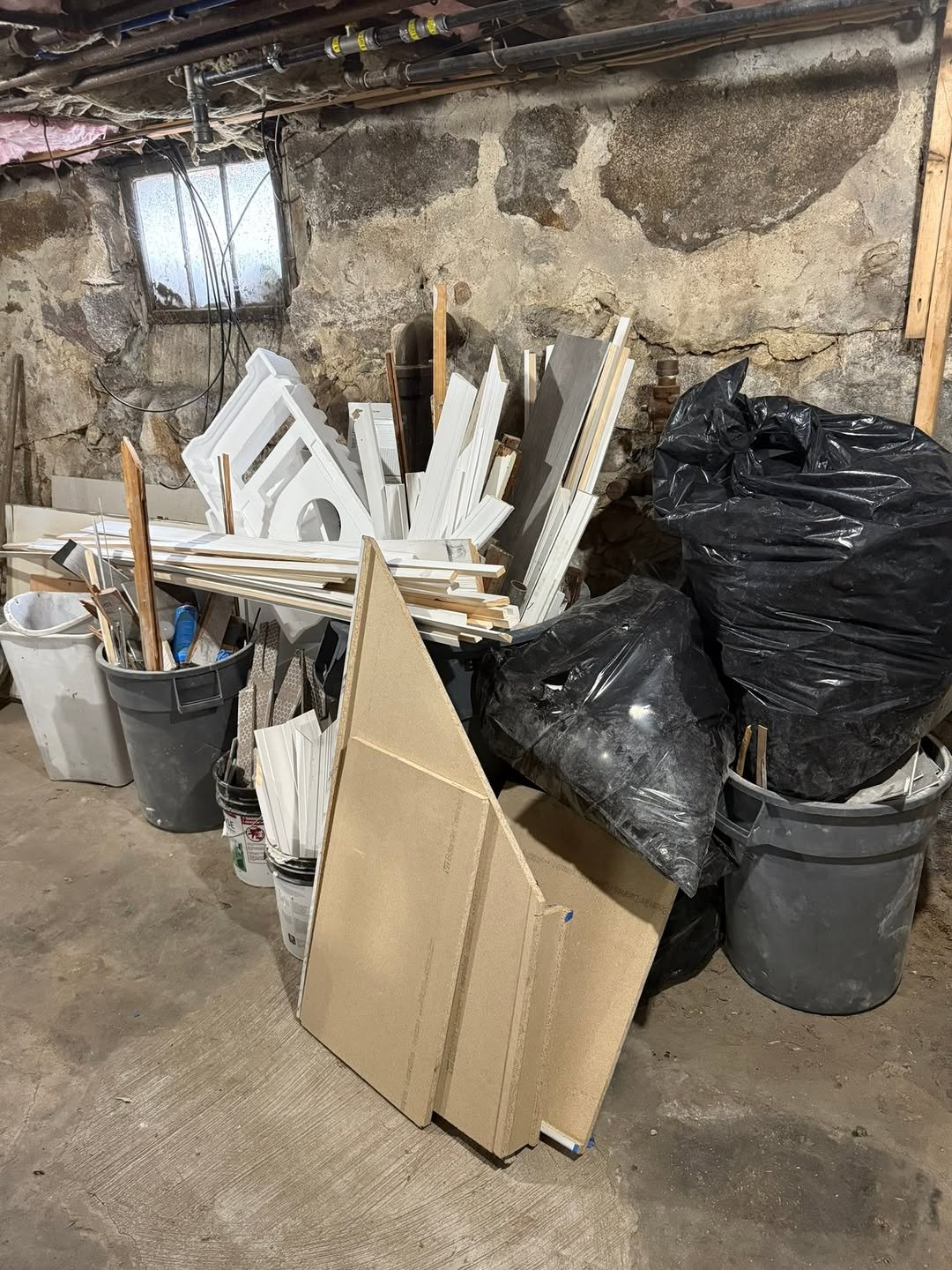 A cluttered workroom with construction supplies, including white and beige wood trim and panels, black garbage bags, buckets, and a small window on a rough stone wall.