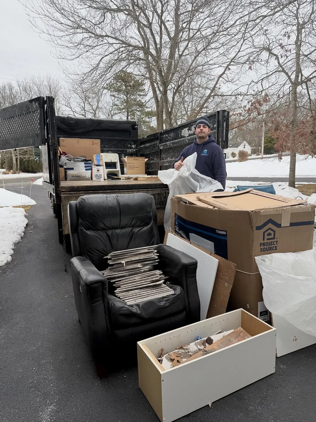 A man with a beard and dark clothing standing next to a truck filled with boxes and supplies on a snowy suburban street.