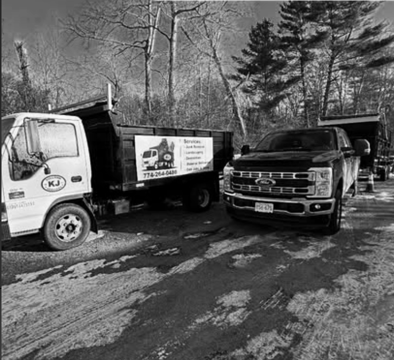Two trucks parked on a muddy, snow-covered lot with trees in the background. One truck has a sign advertising services like auto body and landscaping.