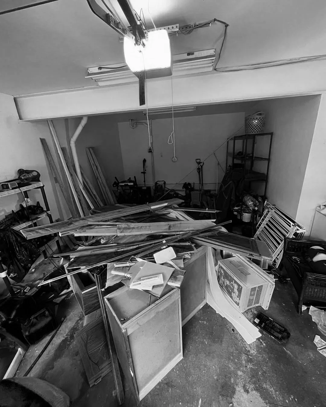 A cluttered workshop with wooden planks, tools, and furniture scattered around, under a bright ceiling light.