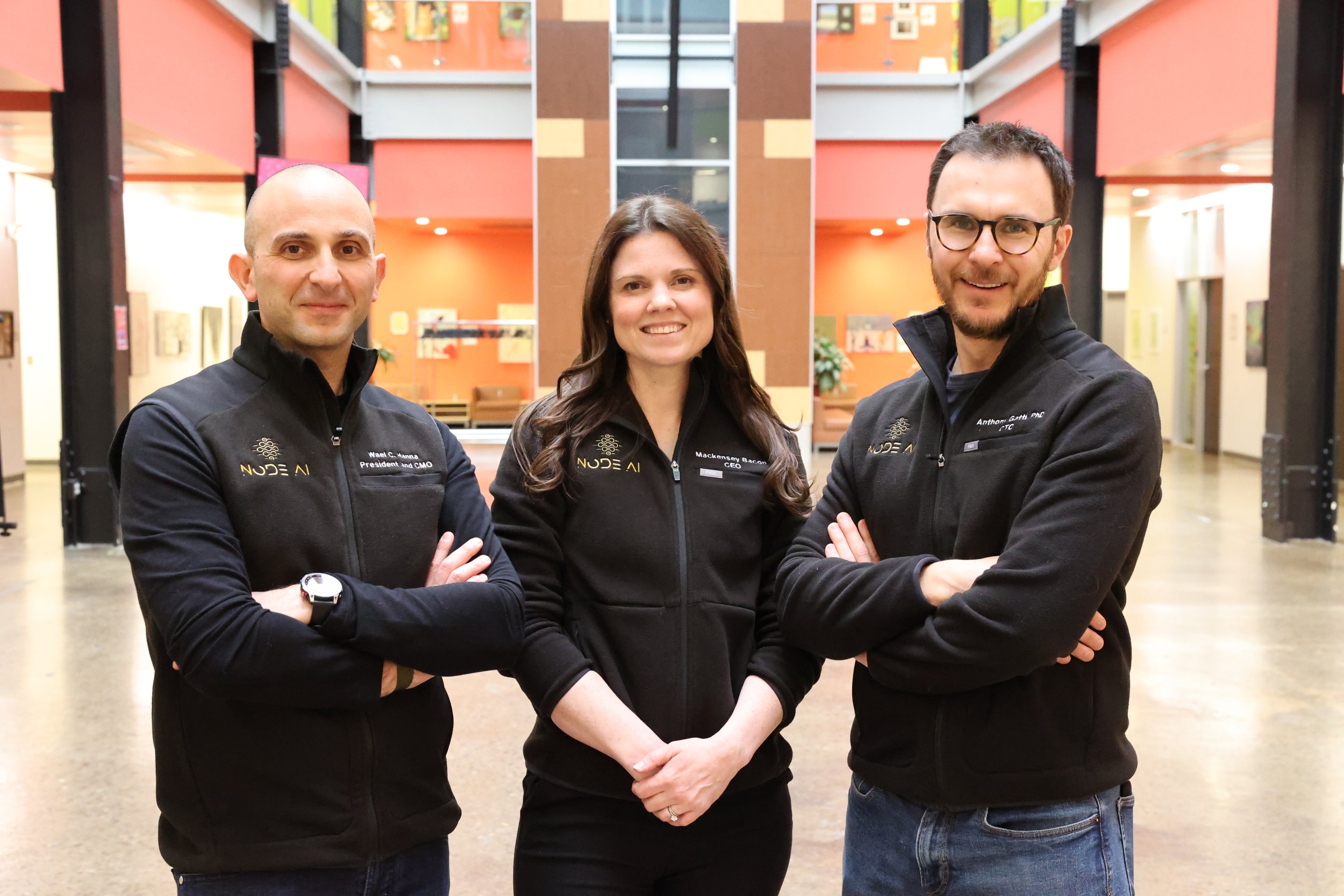Three professionals standing side by side in an office lobby, wearing black jackets with the logo 'NODE AI' and their names and titles on them. Mackensey Bacon, Wael Hanna, Anthony Gatti- Founders of NodeAI.