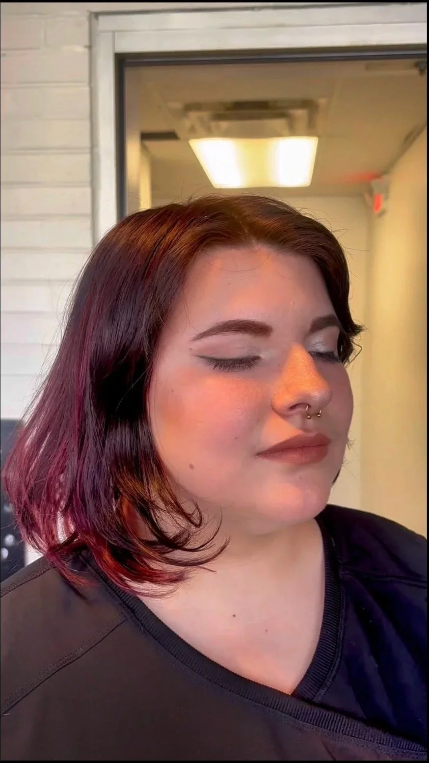 A young woman with closed eyes and dark red hair with purple ends, wearing makeup and a septum piercing, standing indoors.