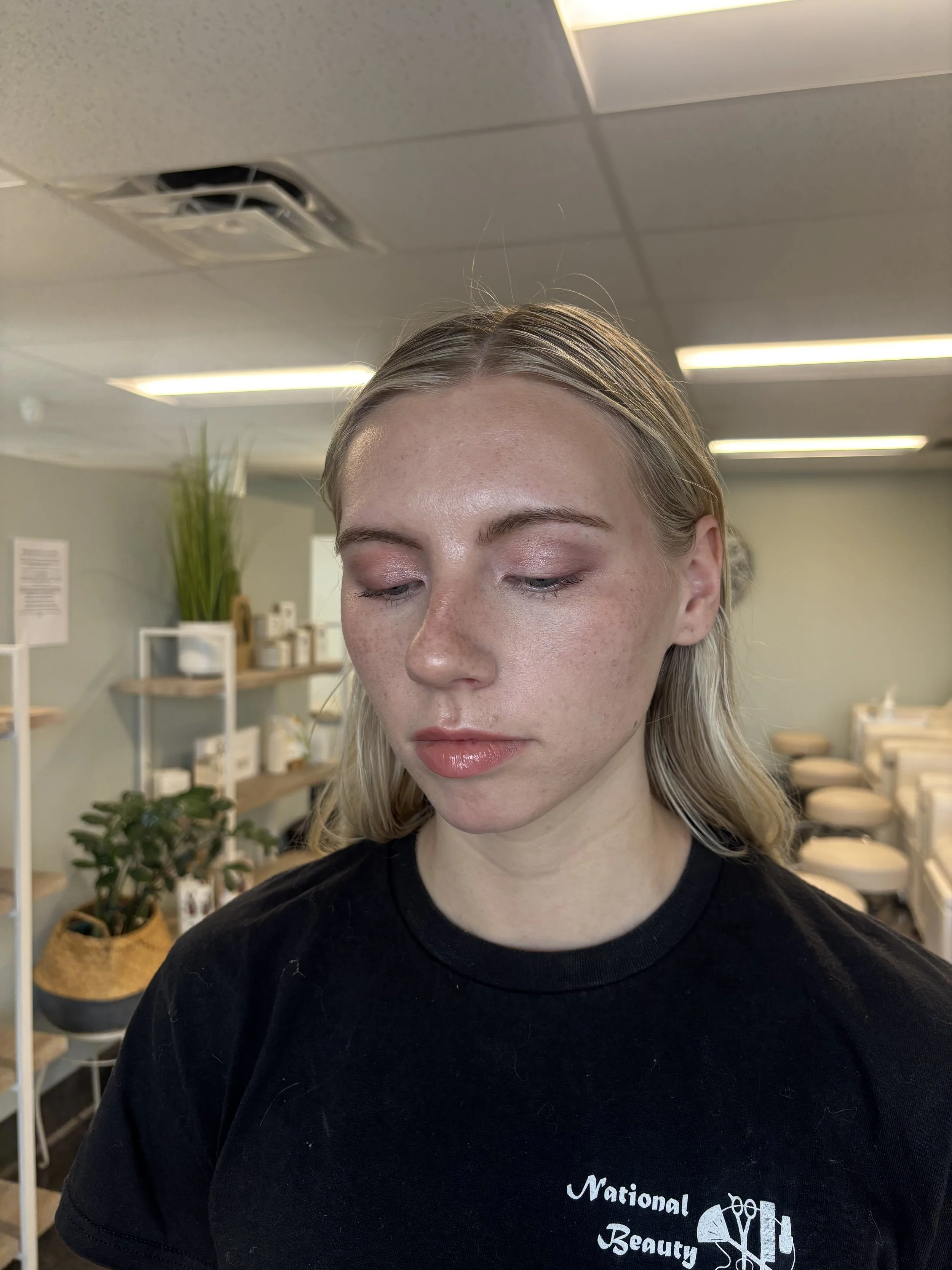 A woman with light blonde hair and natural makeup, wearing a black T-shirt, standing in a beauty or wellness studio with shelves of plants and skincare products in the background.