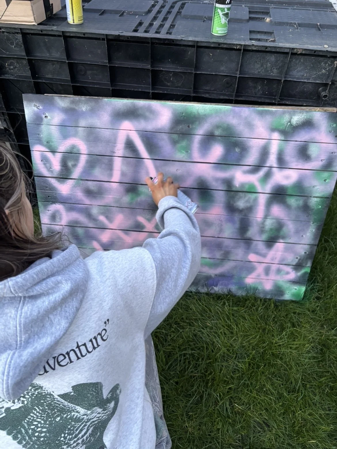 A person wearing a gray sweatshirt is spray painting pink graffiti on a wooden board outdoors. The graffiti includes a heart and some abstract shapes, with some green and black spray paint around.