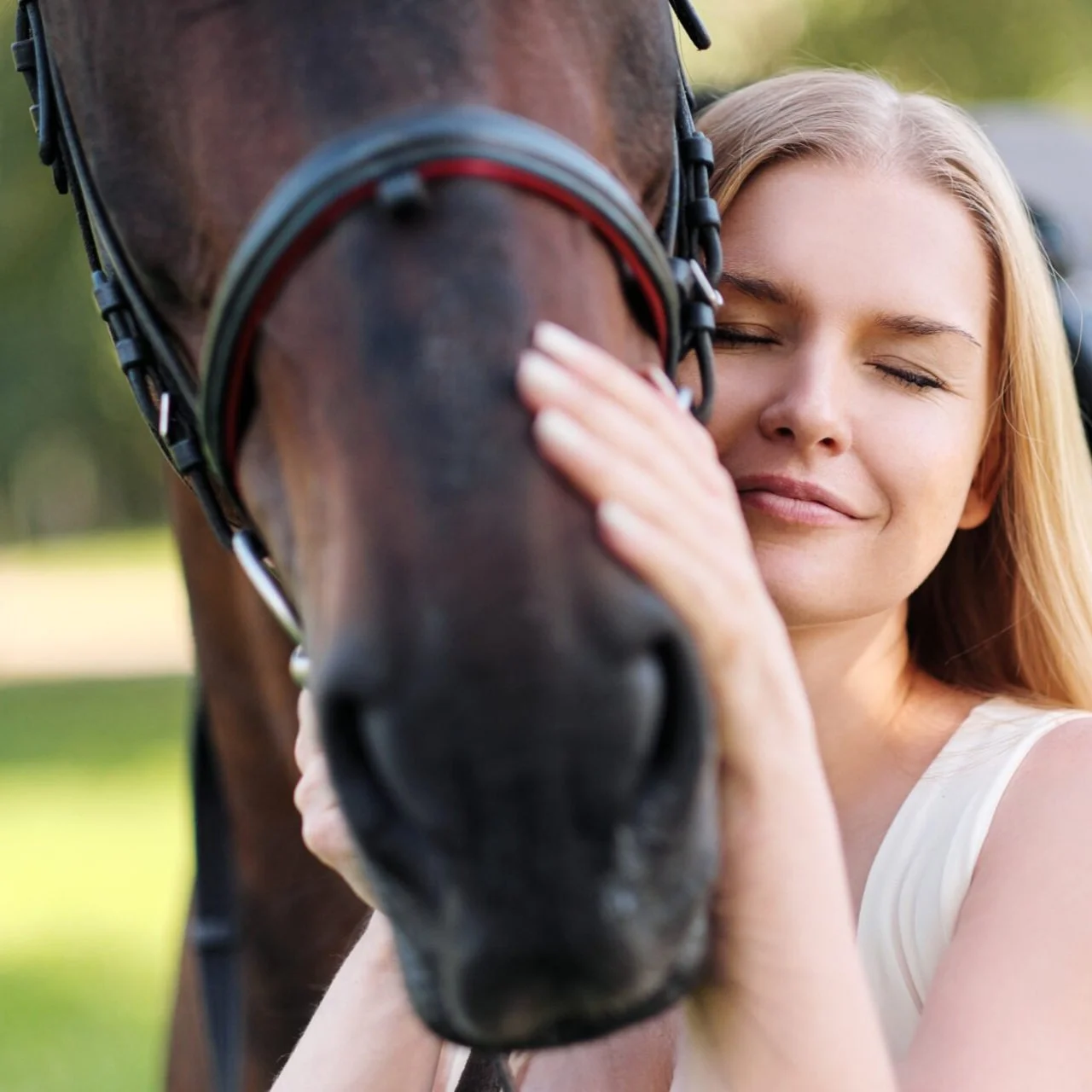 A woman with long blonde hair gently touching the face of a brown horse with a black mane, both with eyes closed, outdoors on a sunny day.