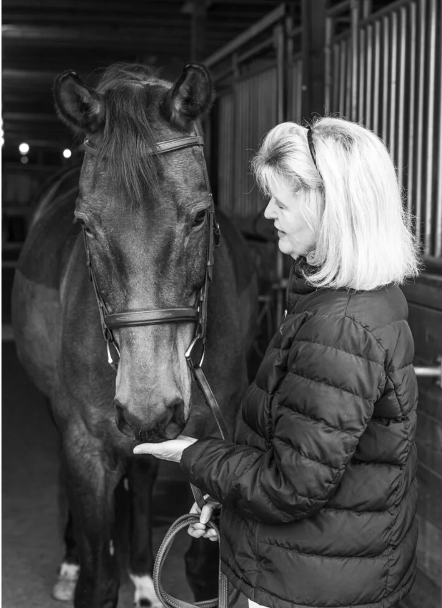 A woman with blonde hair wearing a quilted jacket is gently holding a horse's chin inside a stable, with the horse's head slightly lowered.