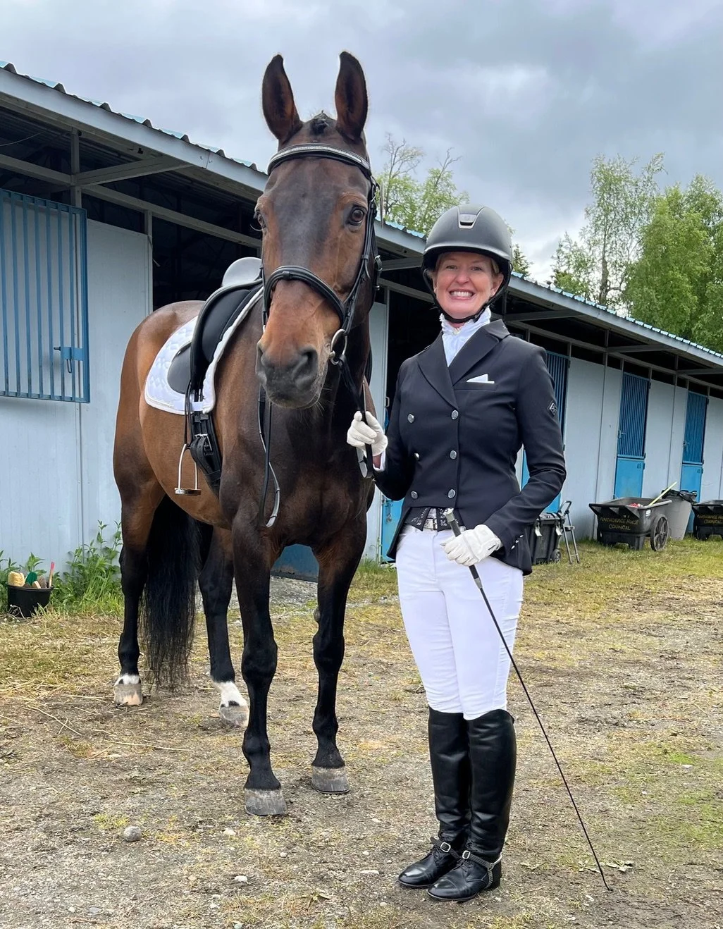 A woman dressed in traditional equestrian attire standing next to a brown horse with a black mane and tail. The woman is holding the horse's bridle and smiling. The background shows a stable with blue doors and overcast sky.