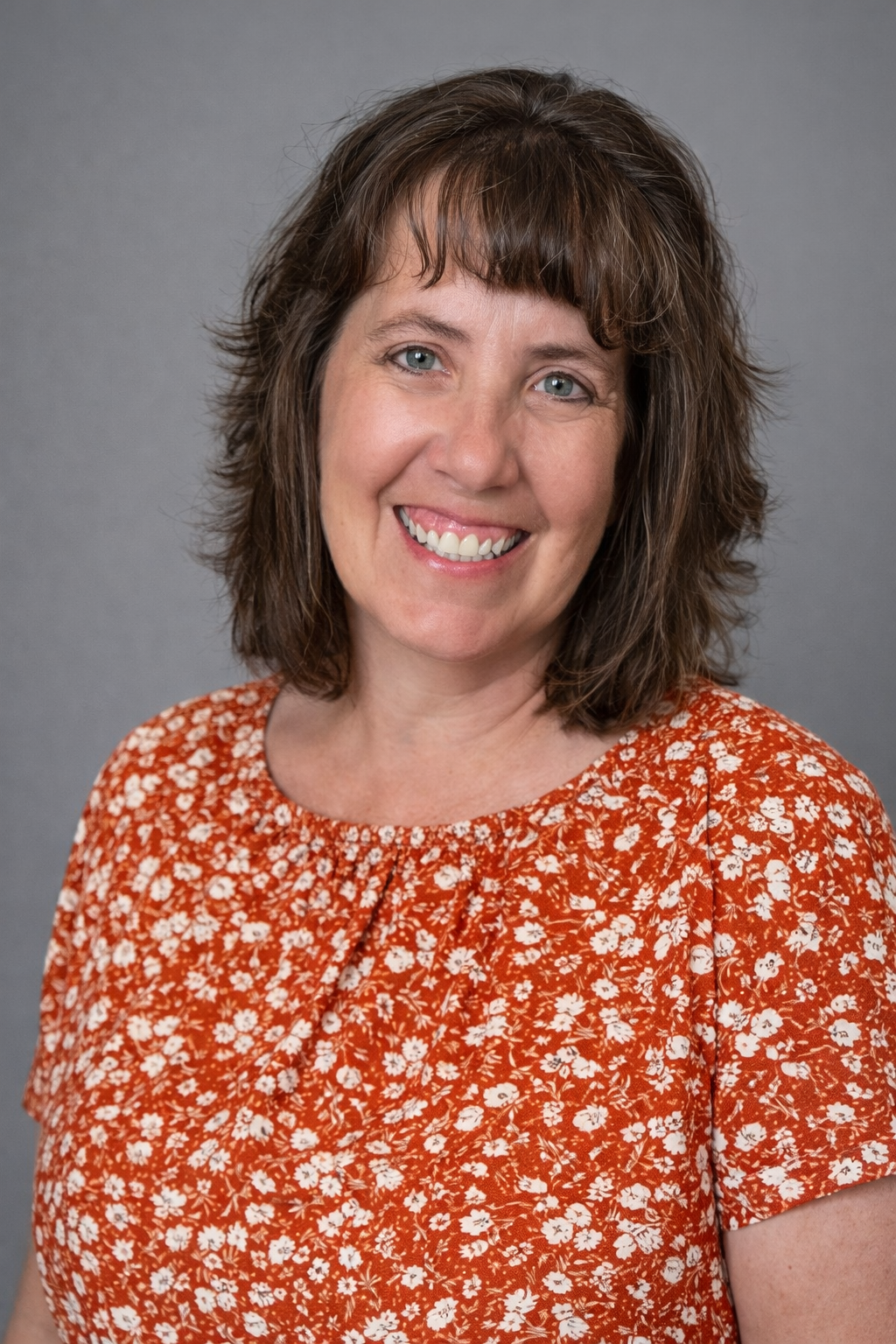 Headshot of a woman with shoulder-length brown hair and blue eyes, smiling, wearing an orange floral blouse, against a gray background.