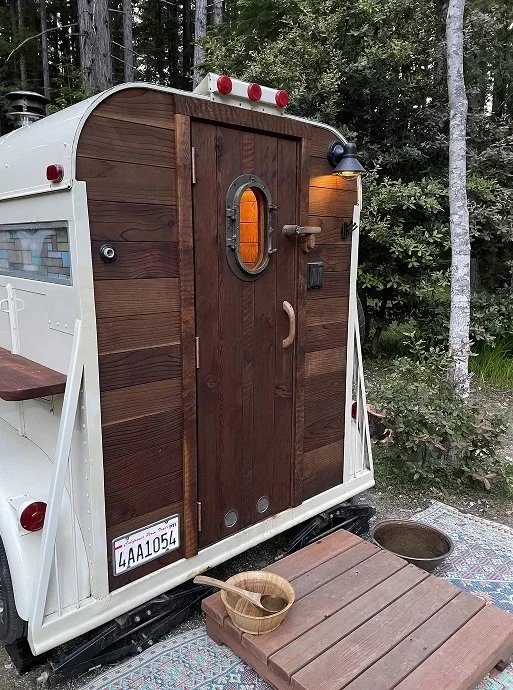 A small camper trailer with a wooden door, mounted on a snowmobile, surrounded by trees. There is a wooden step and bowls in front of the door.
